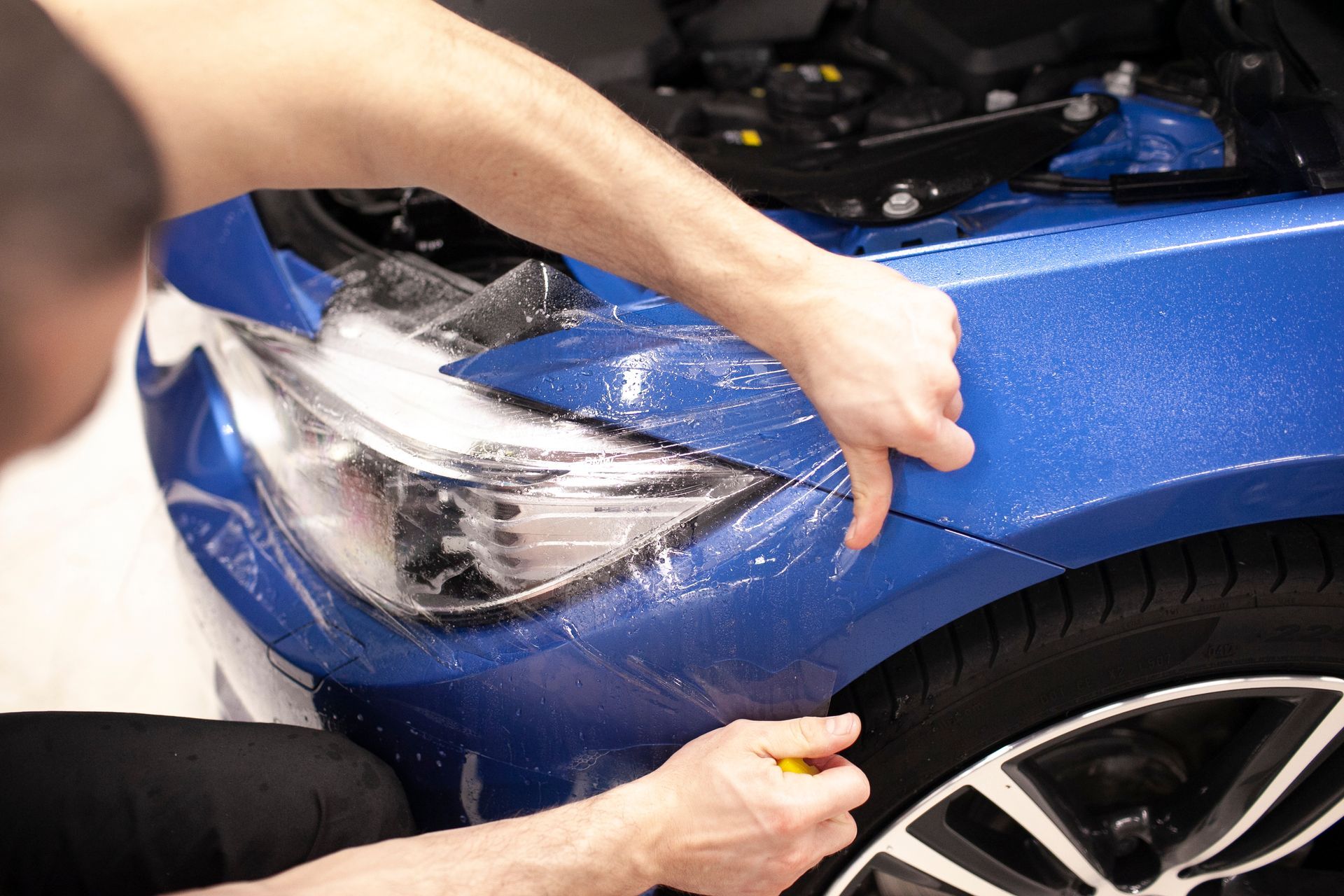 A man is applying a protective film to the front of a blue car.