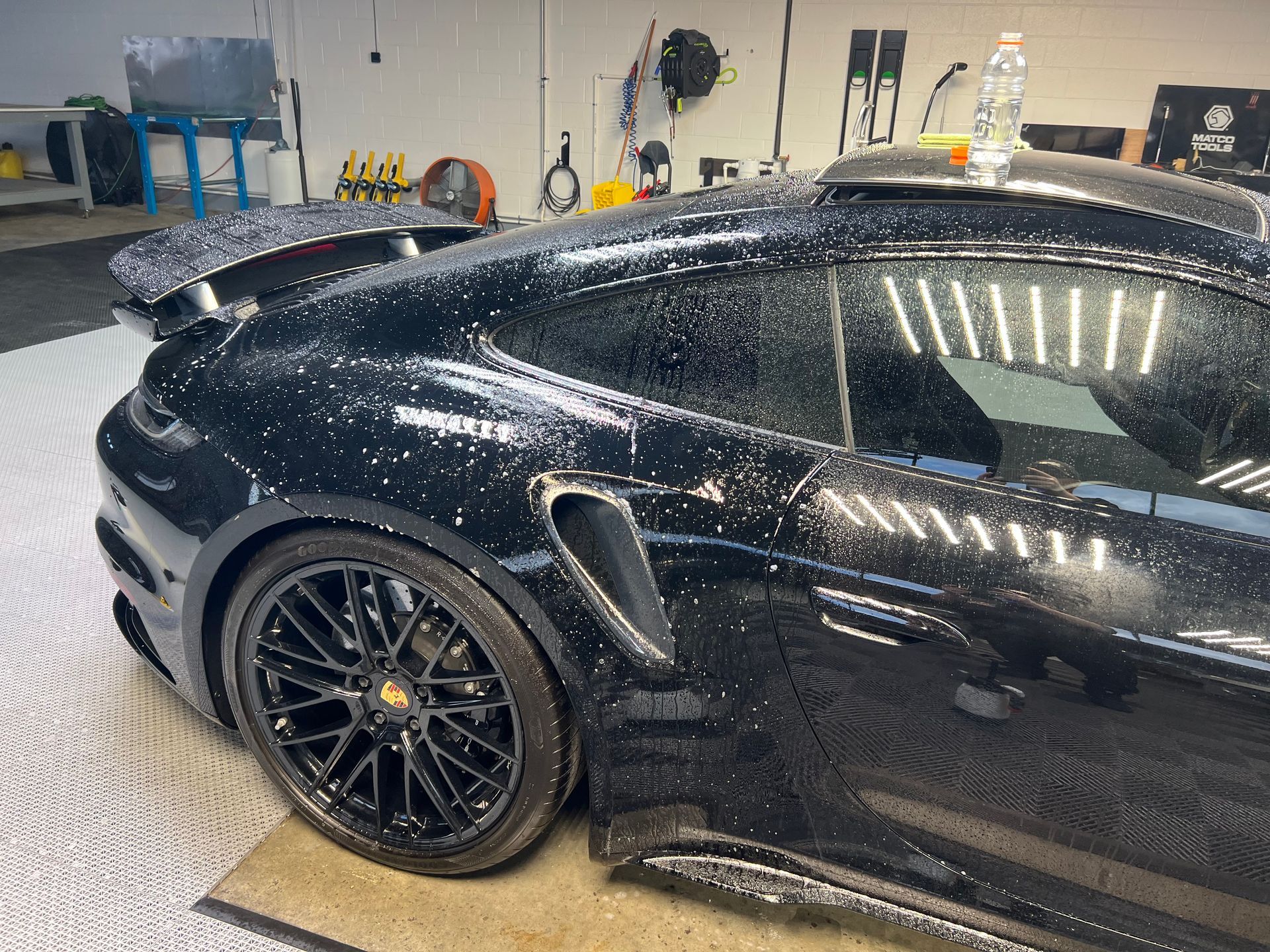 Black Porsche sports car being washed in a garage. Water droplets are visible on the car's surface.
