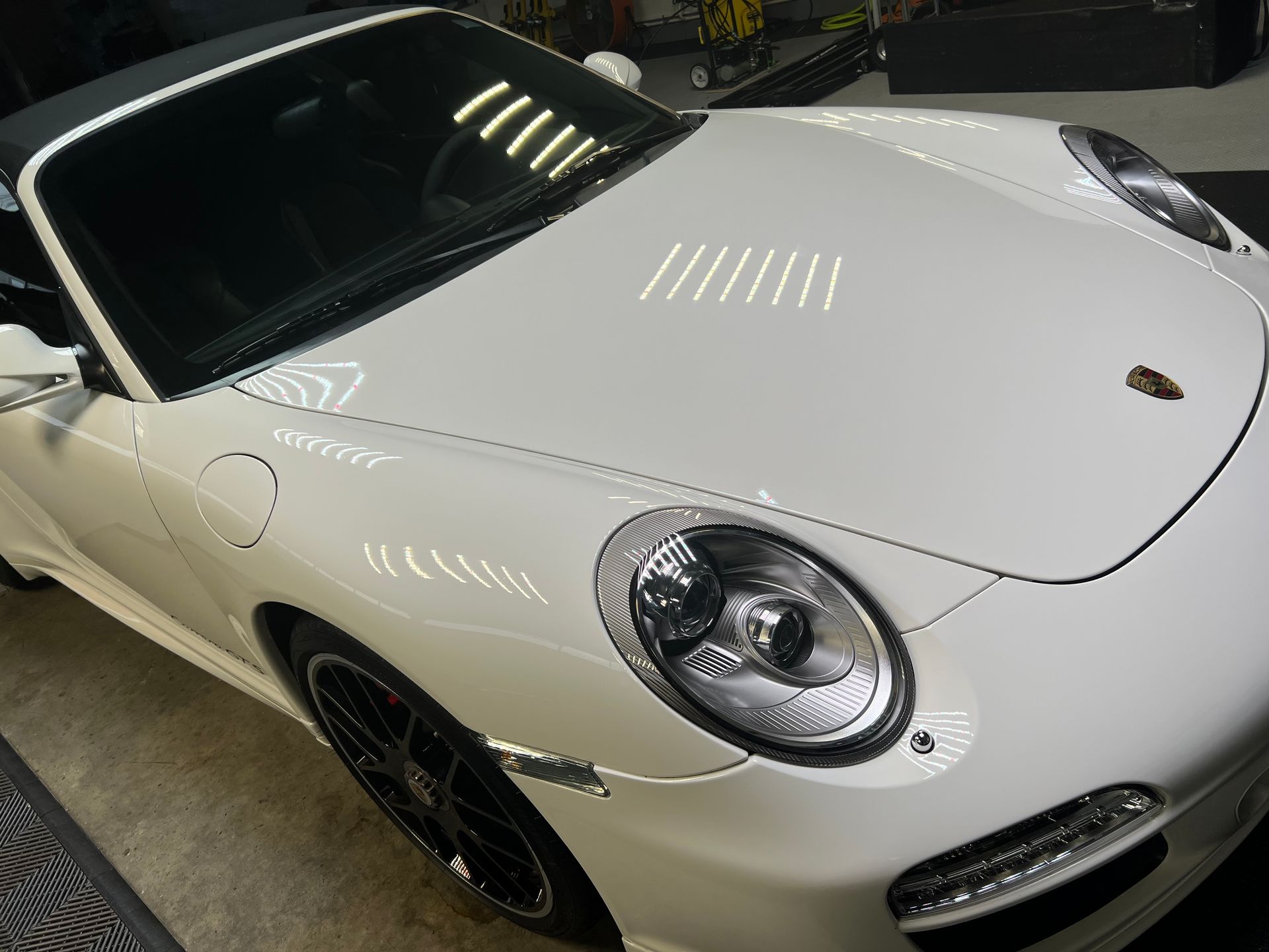 White Porsche convertible, close-up showing the hood, headlights, and black rims, indoors.