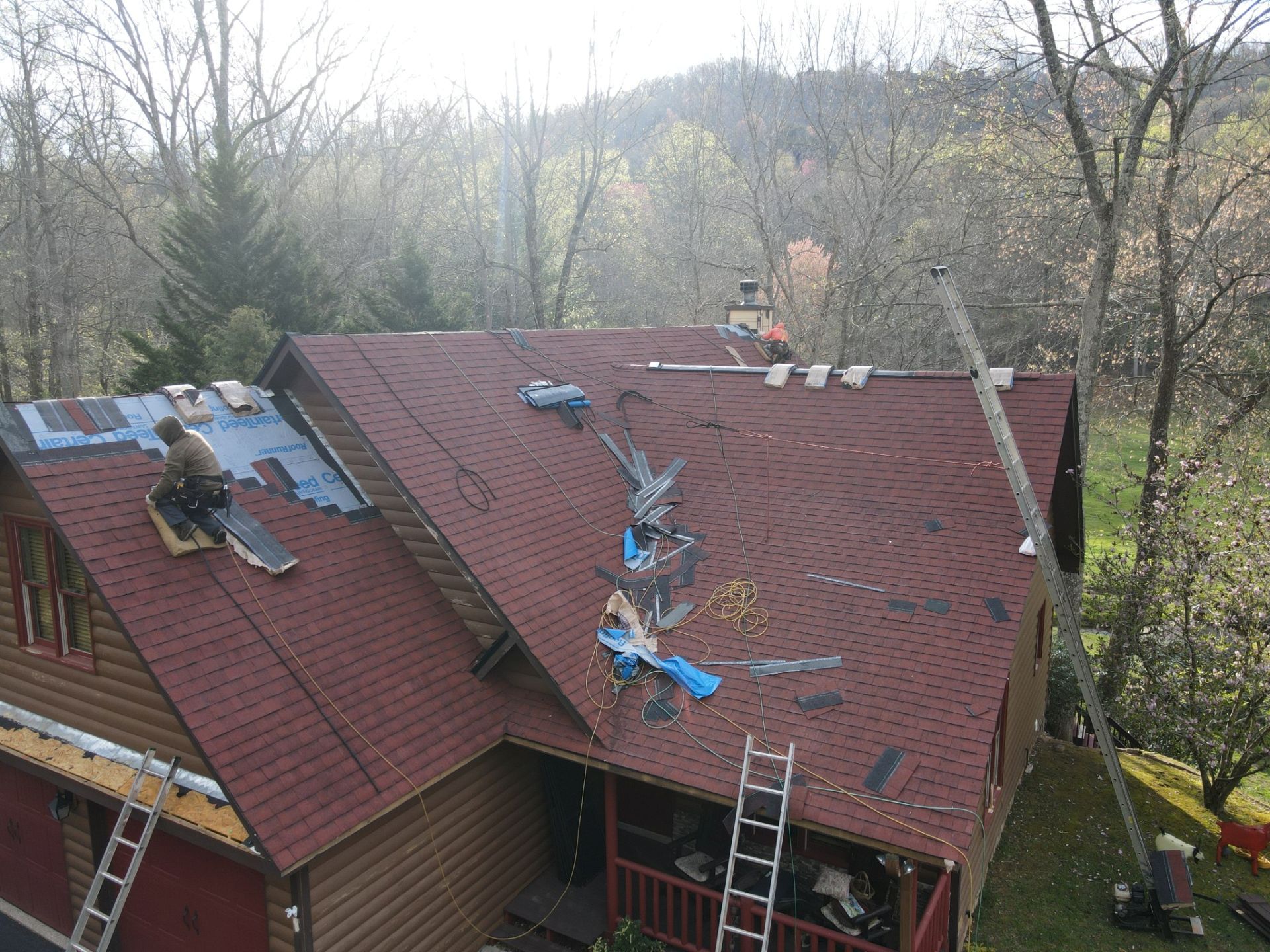 Roofers replacing shingles on a house with a brown roof in a wooded area. Ladders and tools visible.