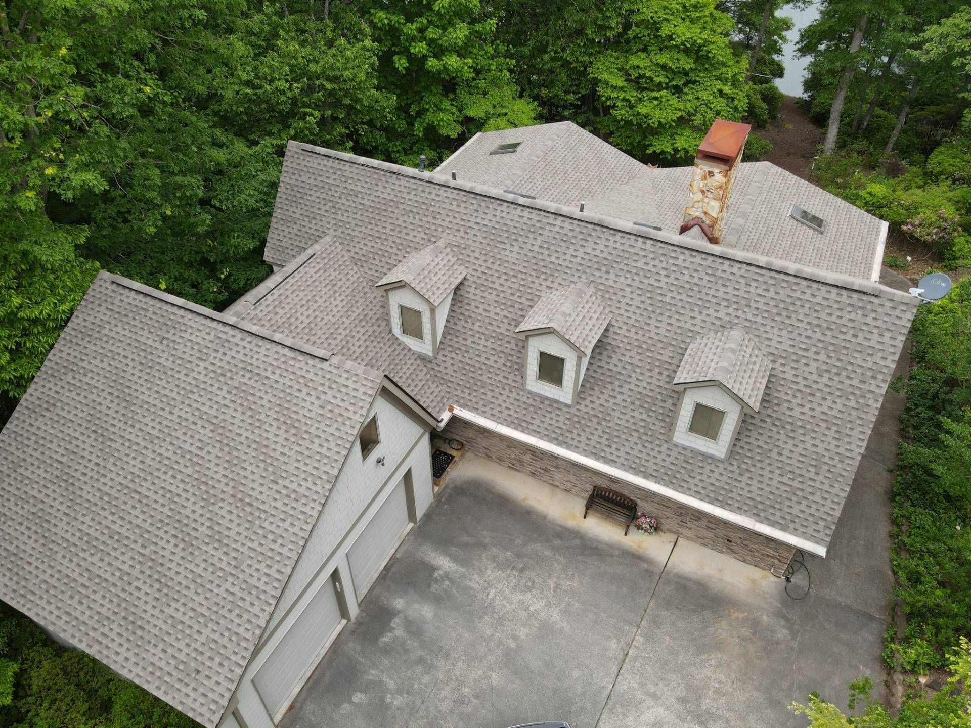 Aerial view of a house with gray shingles, three dormers, and a chimney surrounded by green trees and a paved driveway.