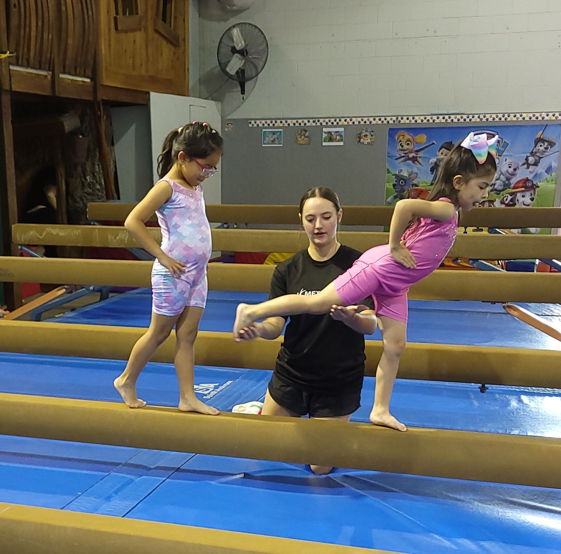 Two young girls practicing gymnastics on balance beams with instructor's help; indoor gym.