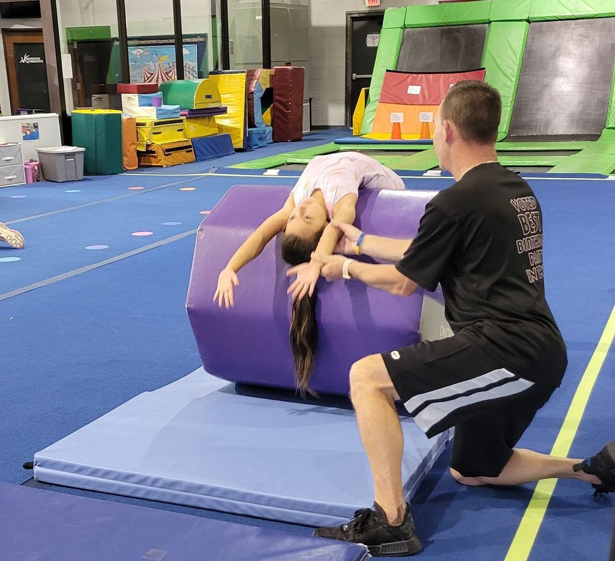 Gymnast doing a back bend on a purple barrel, assisted by a coach. Blue mat, gym setting.