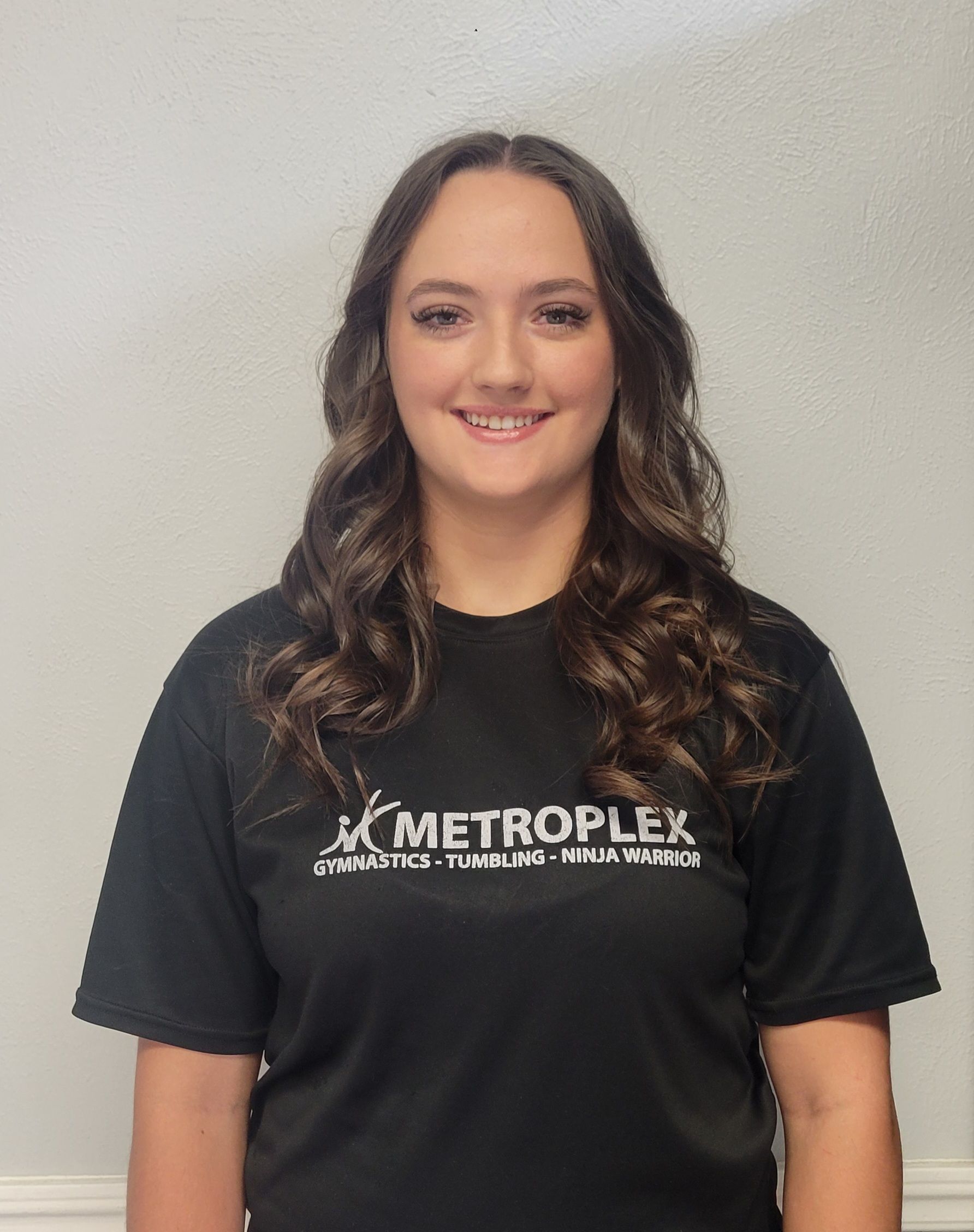 Woman with wavy brown hair, smiling, wearing a black shirt with the Metroplex Gymnastics logo.