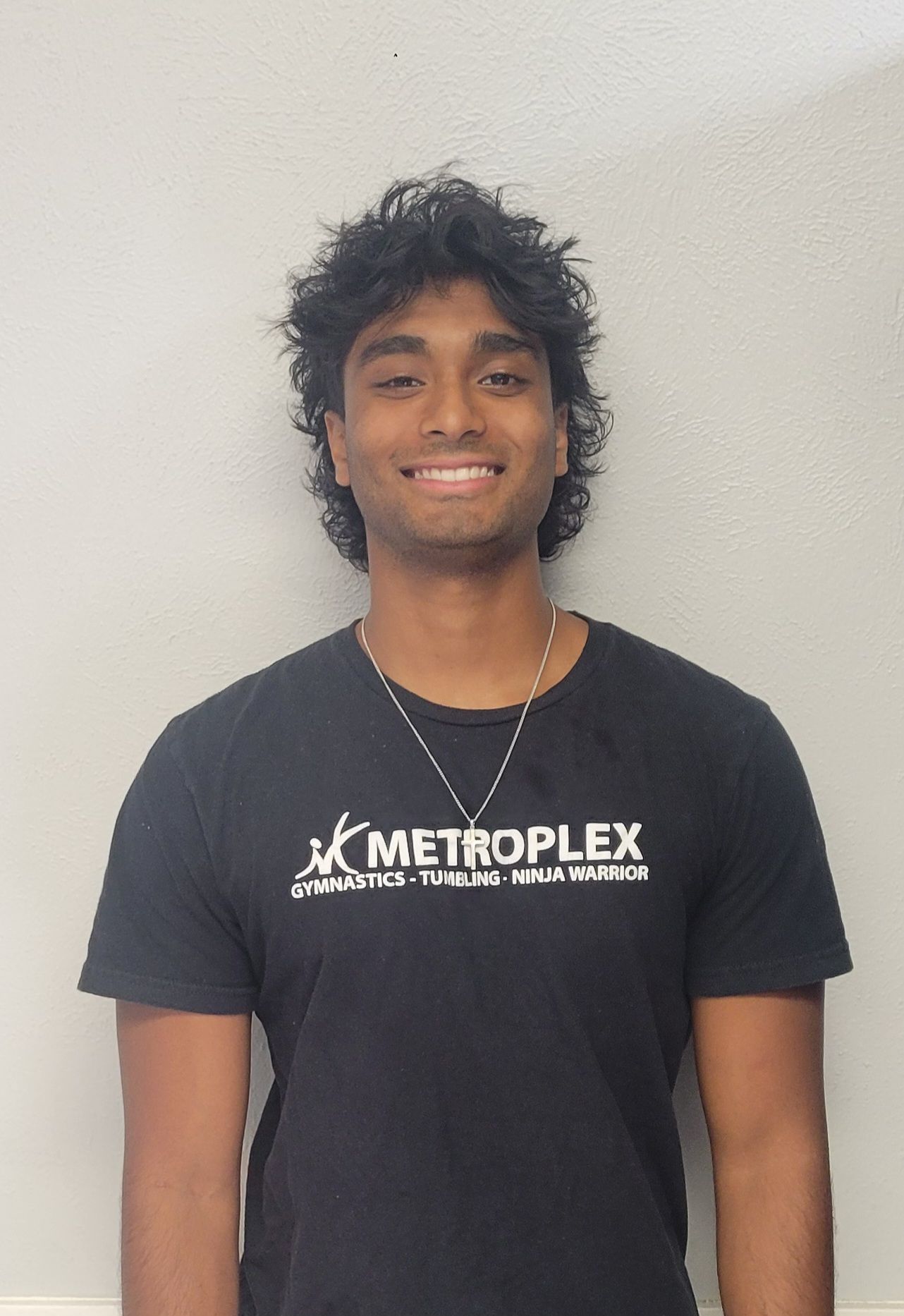 Person smiling, wearing a black t-shirt with a logo, standing in front of a gray wall.