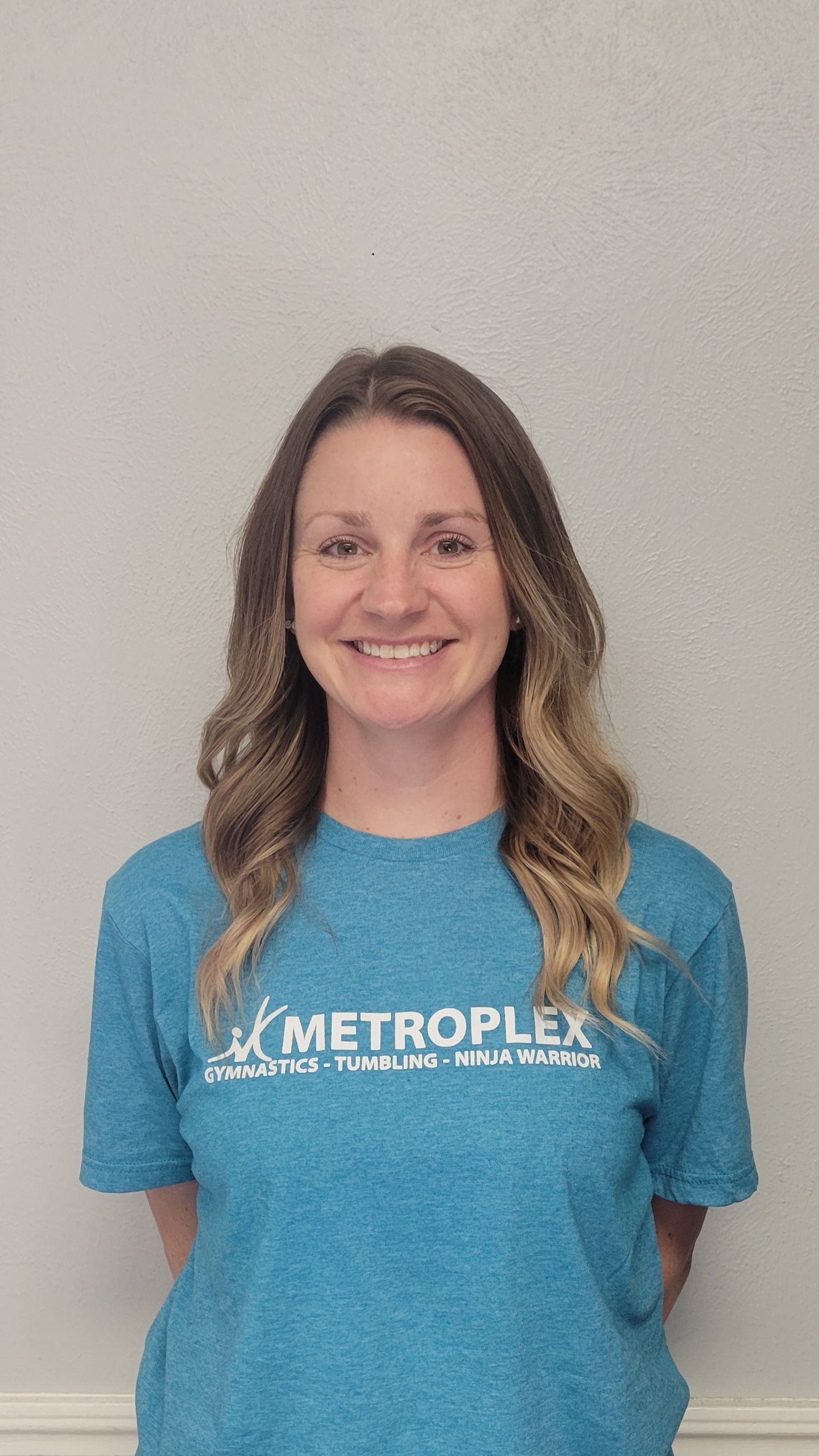 Woman in blue shirt smiles, arms behind back. Text on shirt: 