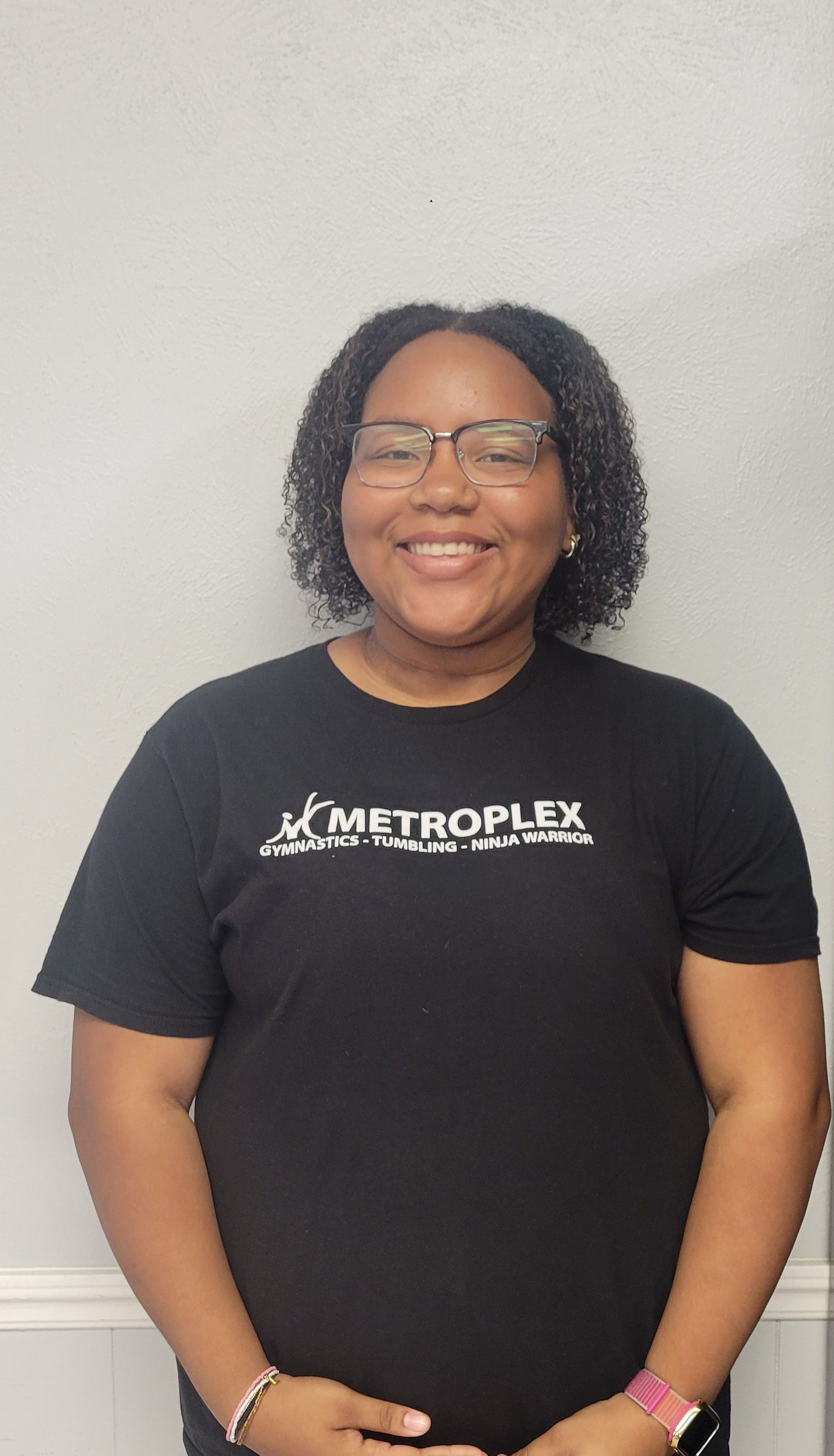 Person wearing glasses smiles in a black shirt with white lettering. Stands in front of a gray wall.