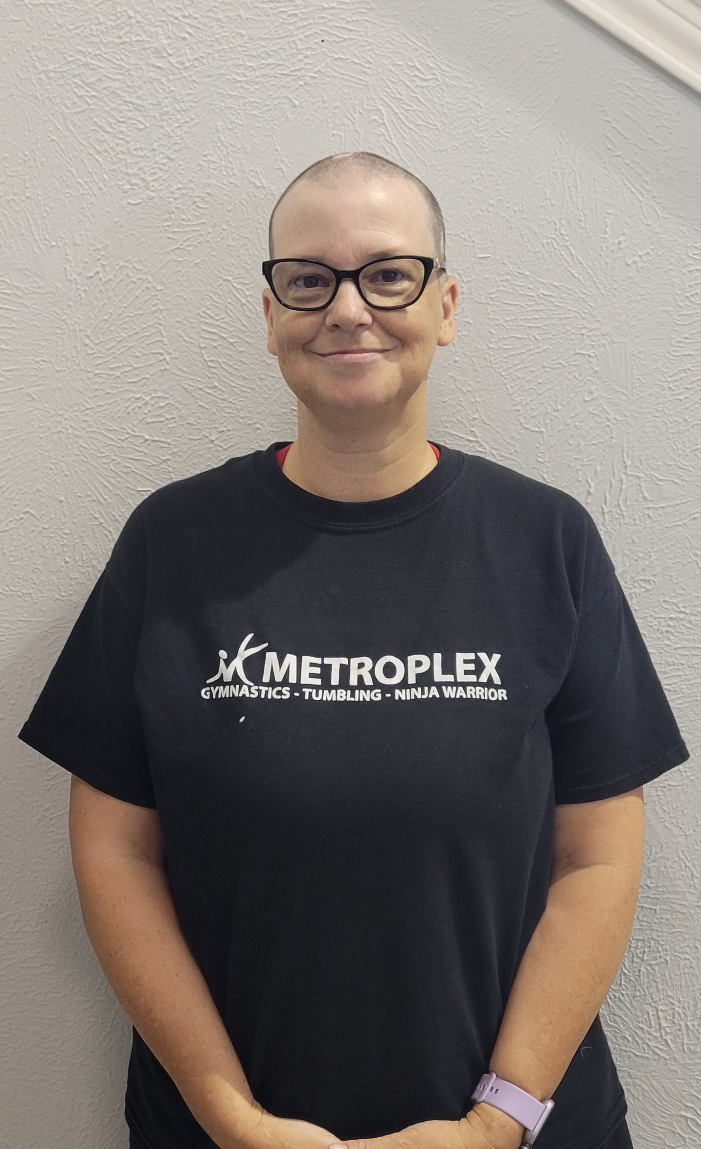 Woman wearing glasses and a black t-shirt with a logo, standing in front of a neutral-colored wall.