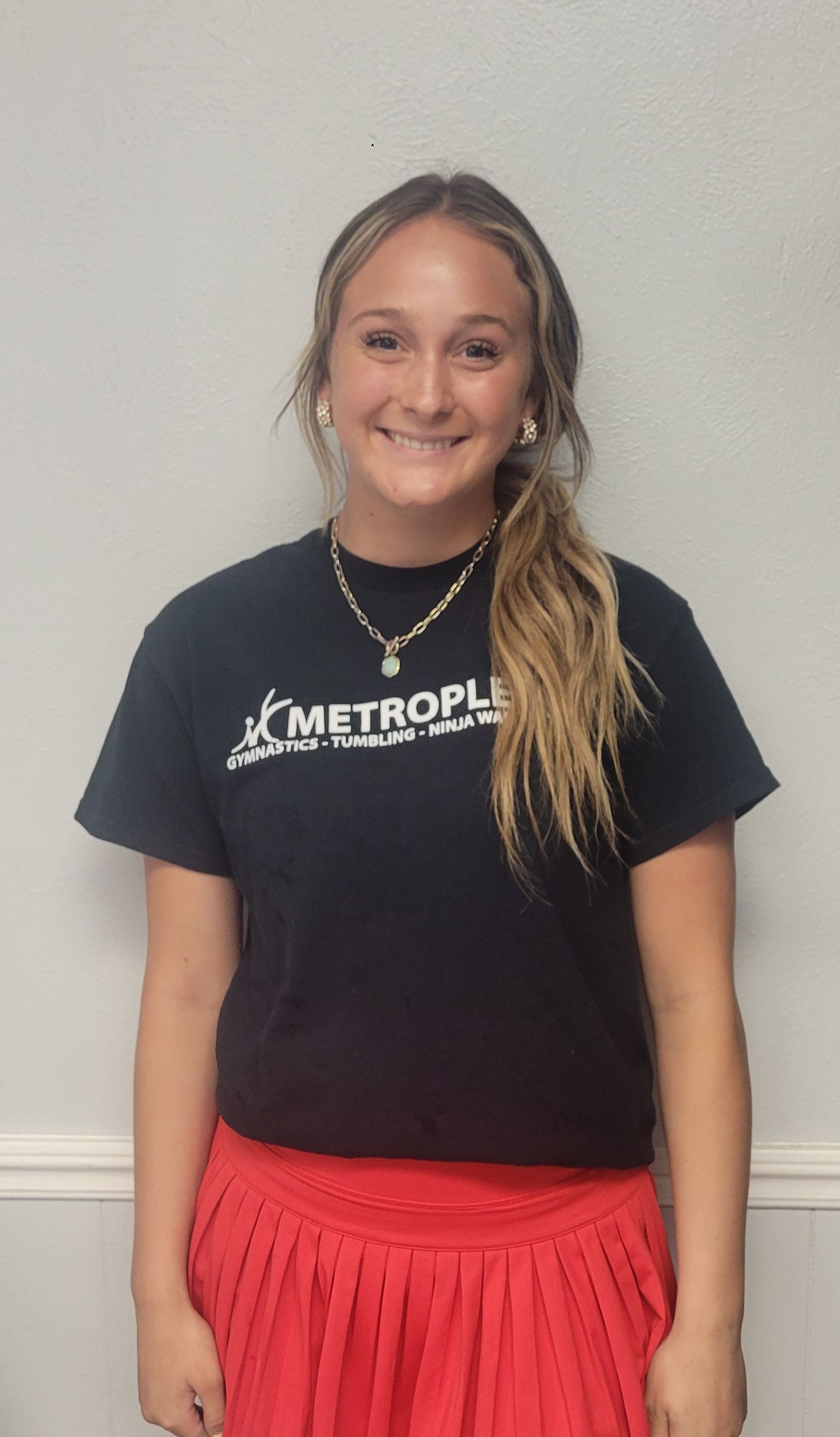 Woman in black t-shirt and red skirt smiles at camera, standing in front of a white wall.