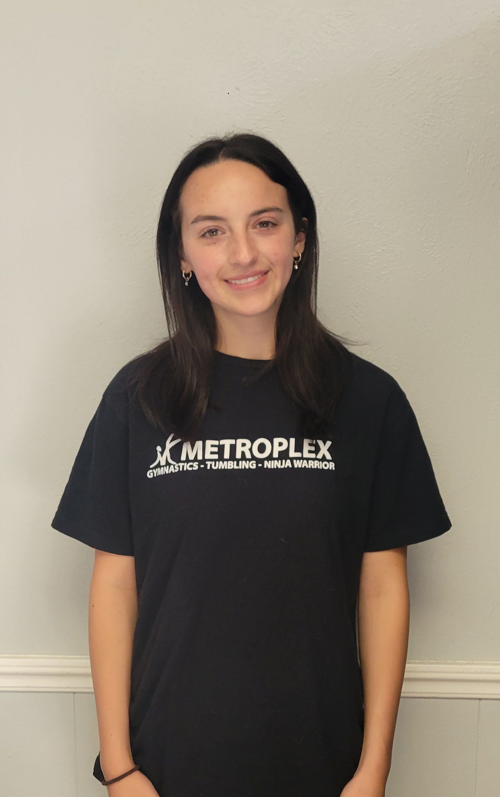 Young person in a black shirt with text, smiling, standing in front of a neutral wall.
