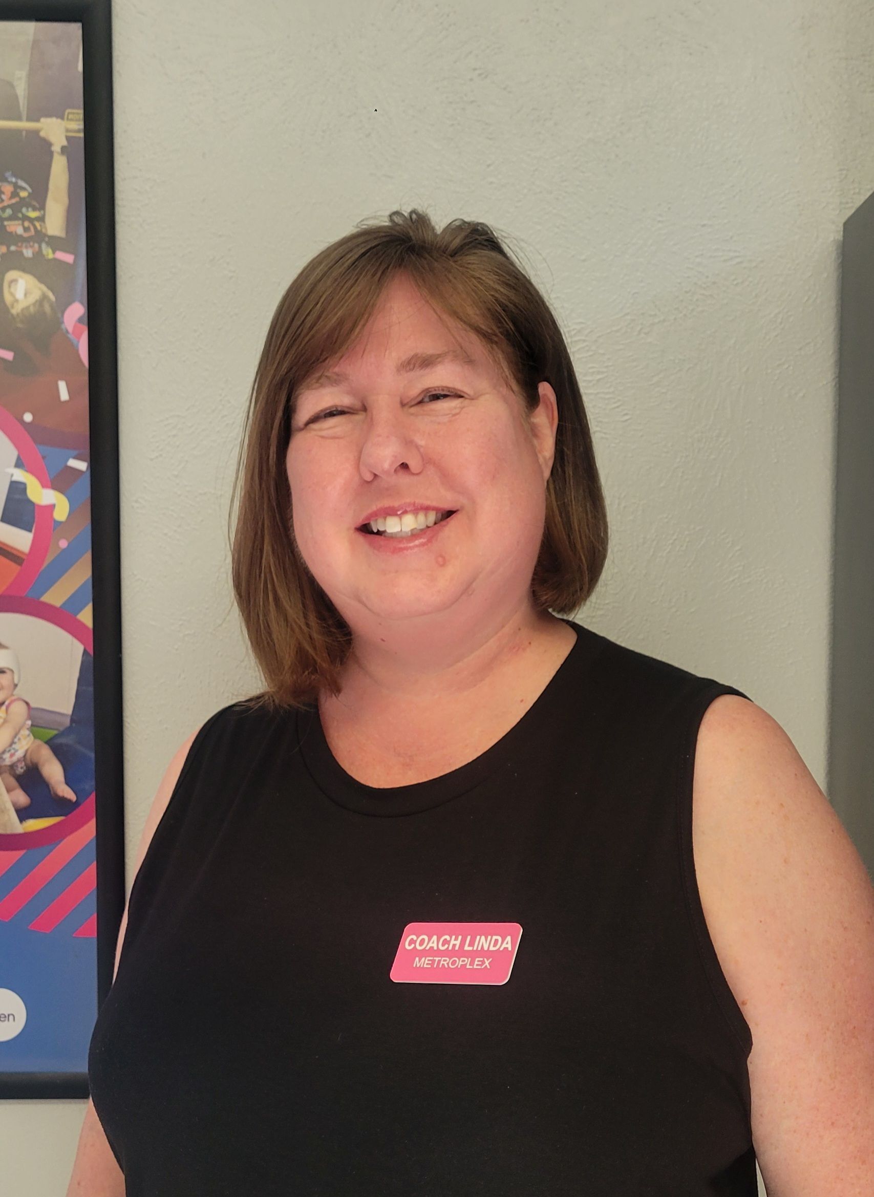 Woman with short brown hair, smiling, wearing a black top. There's a pink logo on her shirt.