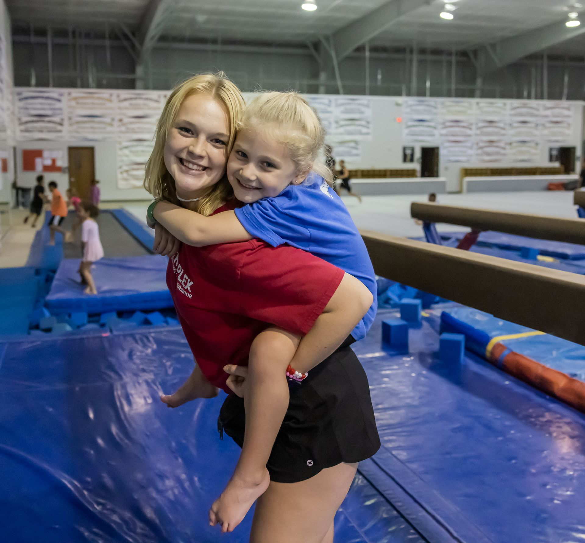 Two girls in a gym. One gives the other a piggyback ride. Both smile. Blue mats and balance beams in background.