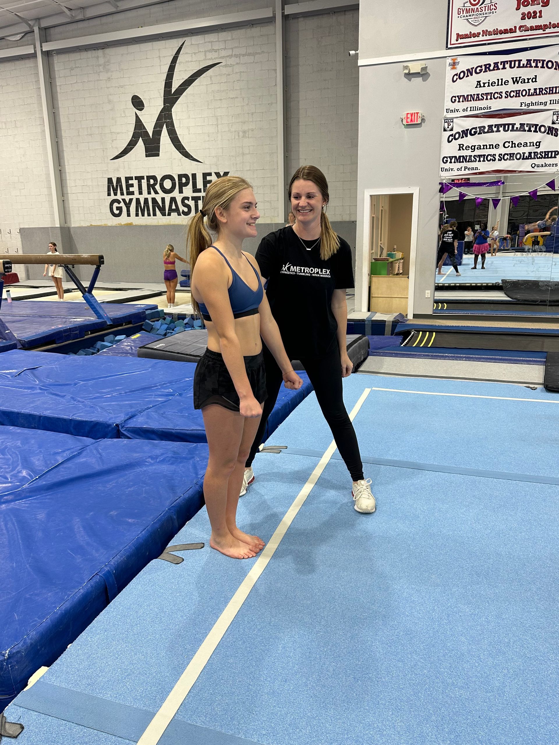 Gymnast and coach on blue mat in a gym. The coach smiles as the gymnast stands ready, smiling too.