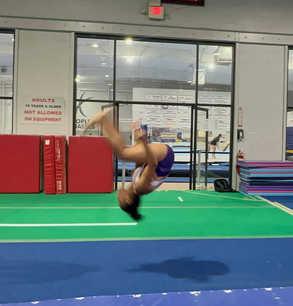 Gymnast in purple and white, mid-air doing a backflip on a blue and green mat inside a gym.