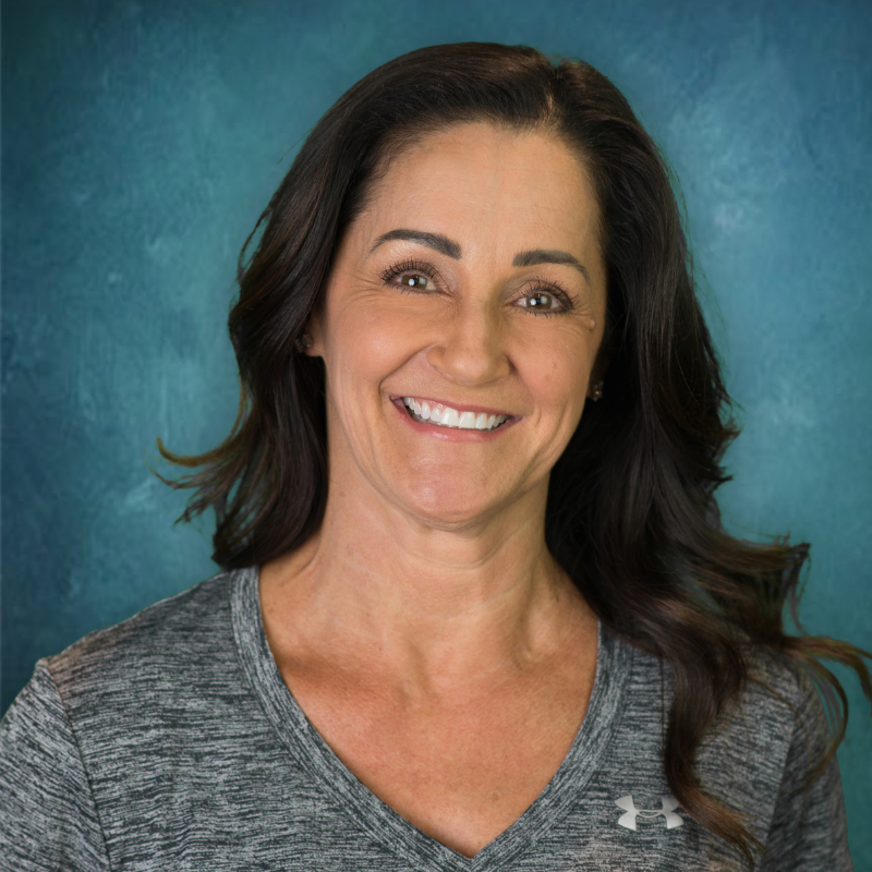 Woman with dark hair smiles, wearing a grey Under Armour shirt against a teal backdrop.