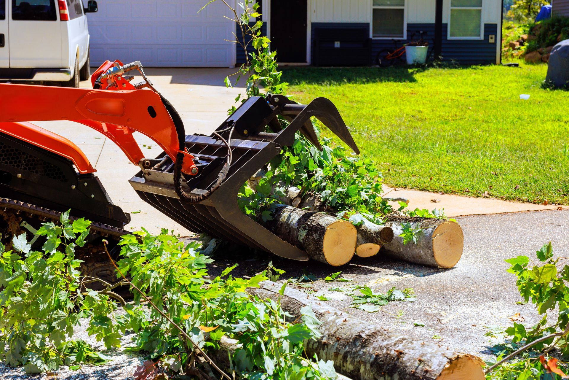 Orange skid steer with grapple bucket lifting logs on a driveway, near a house.