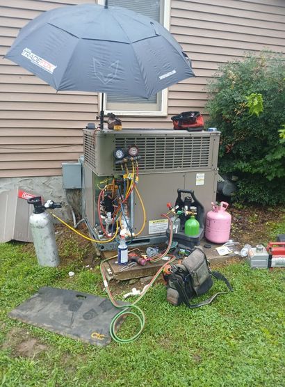 Outdoor HVAC repair setup with equipment, hoses, and tools under a gray umbrella beside a house