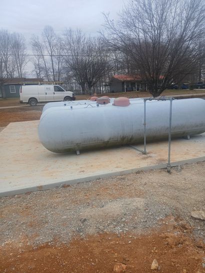 Large gray propane tank on a gravel lot with a white van and bare trees in the background