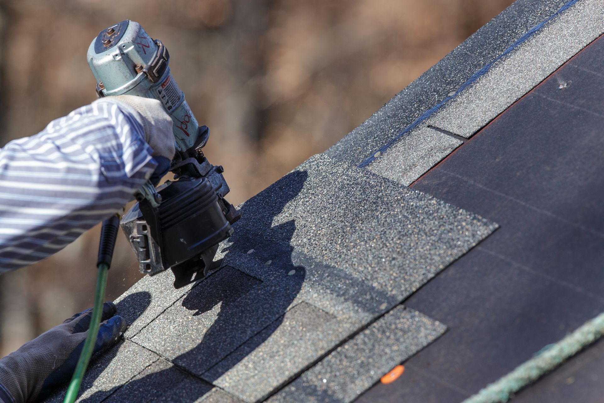 A worker secures asphalt shingles with a nail gun while installing materials on a sloped roof.