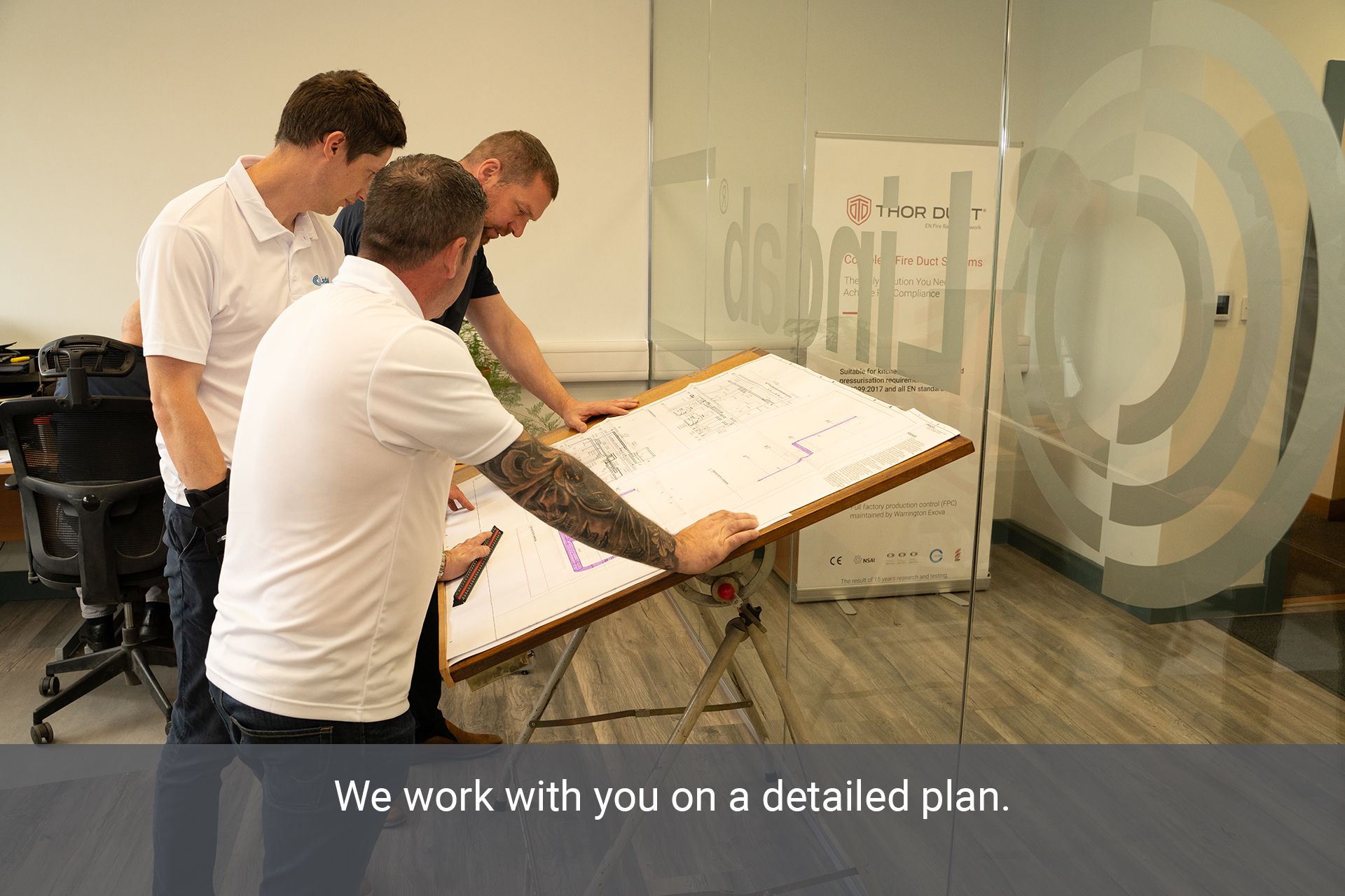 Three A.C. Manufacturing staff in an office looking a drawings on a table with the Lindab logo on a glass wall