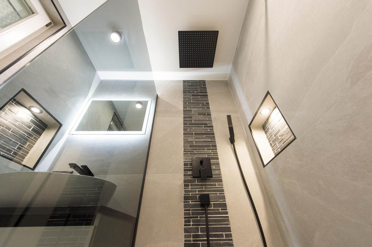 Modernes Badezimmer mit dunklem Duschkopf, eingebautem Regal, beleuchtetem Spiegel und grau-weißen Fliesen.