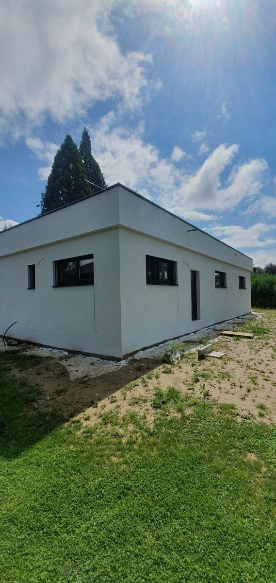 Weißes, rechteckiges Gebäude mit dunklen Fenstern und Flachdach. Heller, sonniger Tag mit blauem Himmel.