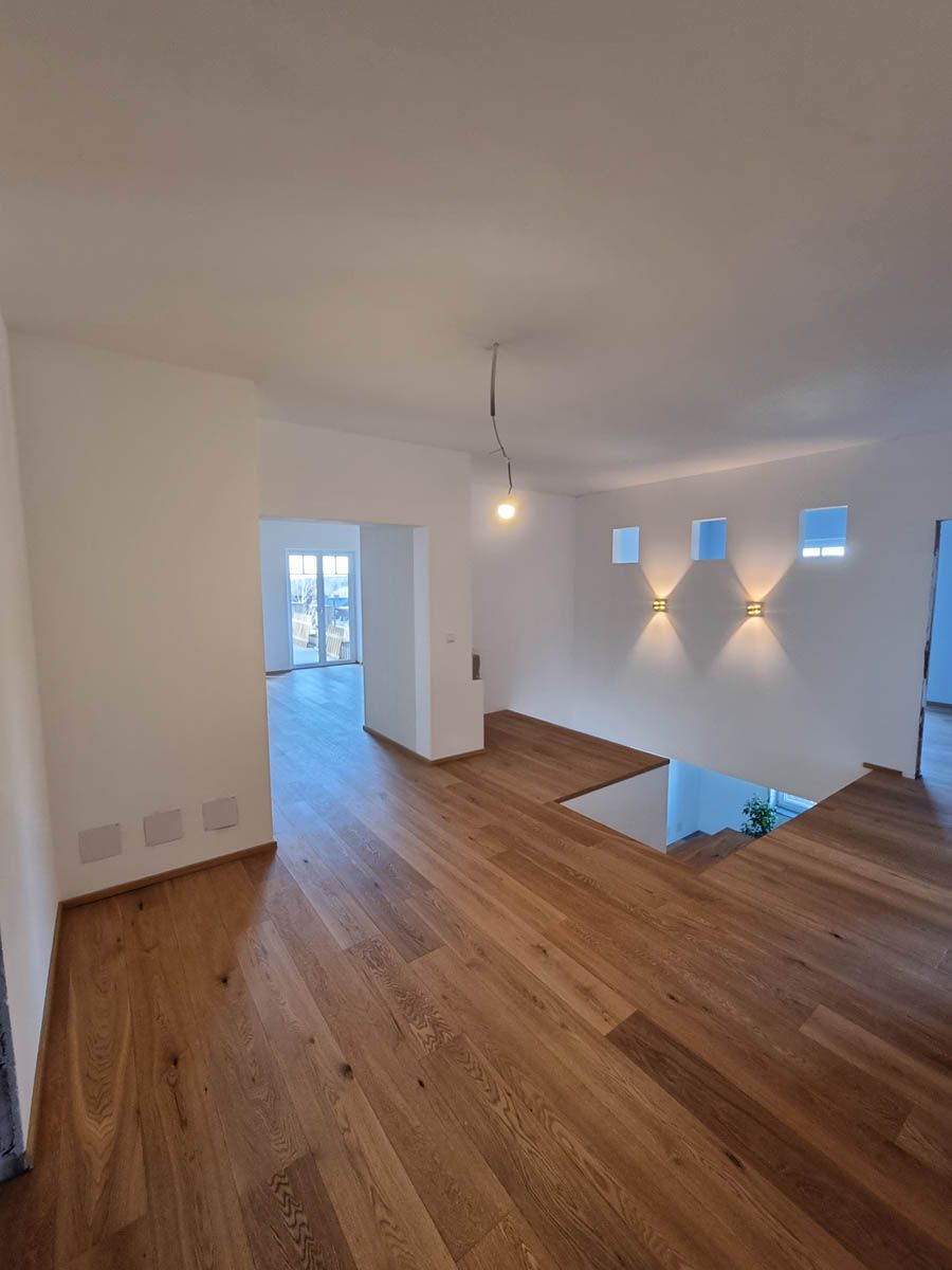 Modernes Interieur mit Holzboden, weißen Wänden, Einbauleuchten, offener Türrahmen, teilweiser Blick auf die Treppe.