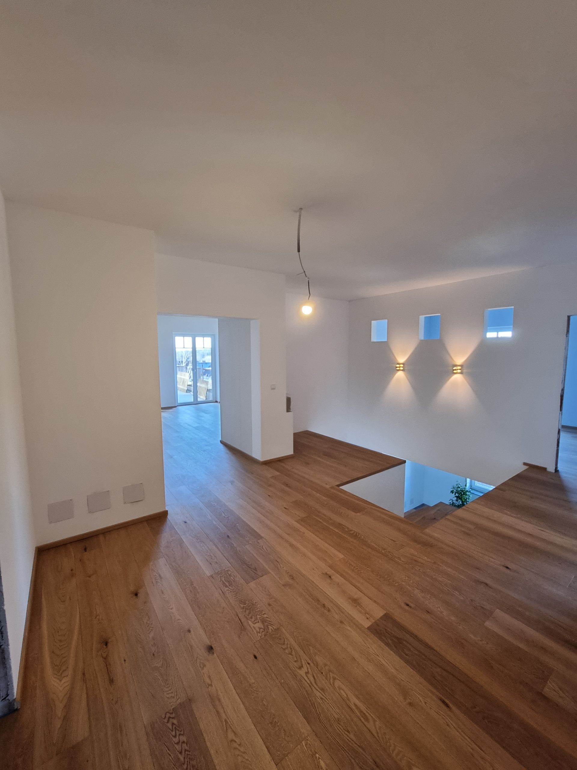 Offenes Interieur mit Holzböden und einer Treppe, die in eine untere Ebene führt. Weiße Wände und Akzentbeleuchtung.