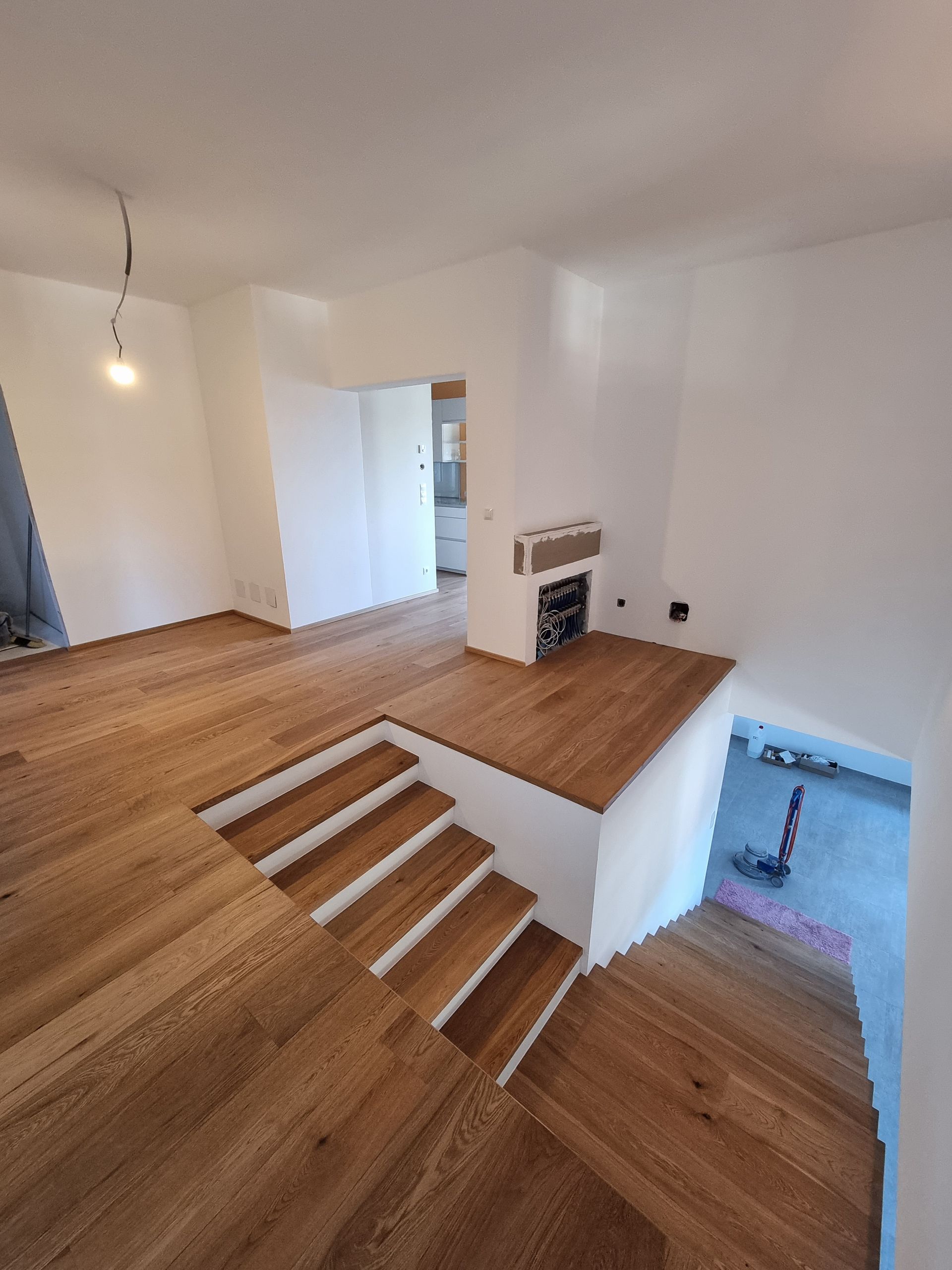 Holzboden mit einer kleinen Treppe, die zu einer erhöhten Plattform mit Kamin führt; in der Ferne ist ein Türrahmen sichtbar.