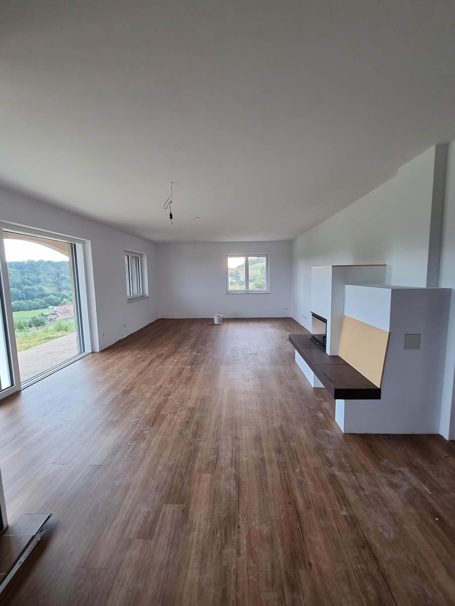 Leeres, helles Wohnzimmer mit Holzboden, weißen Wänden und einem Kamin mit Sitzbank. Große Fenster bieten eine malerische Aussicht.