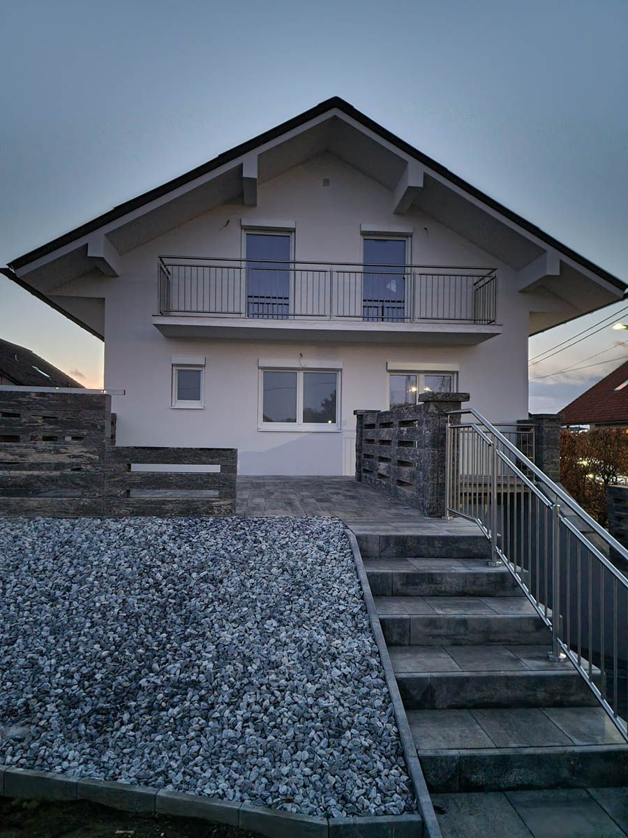 Weißes Haus mit grauem Dach, Balkon und Treppe zum Eingang. Kiesweg und Steinstützmauer.