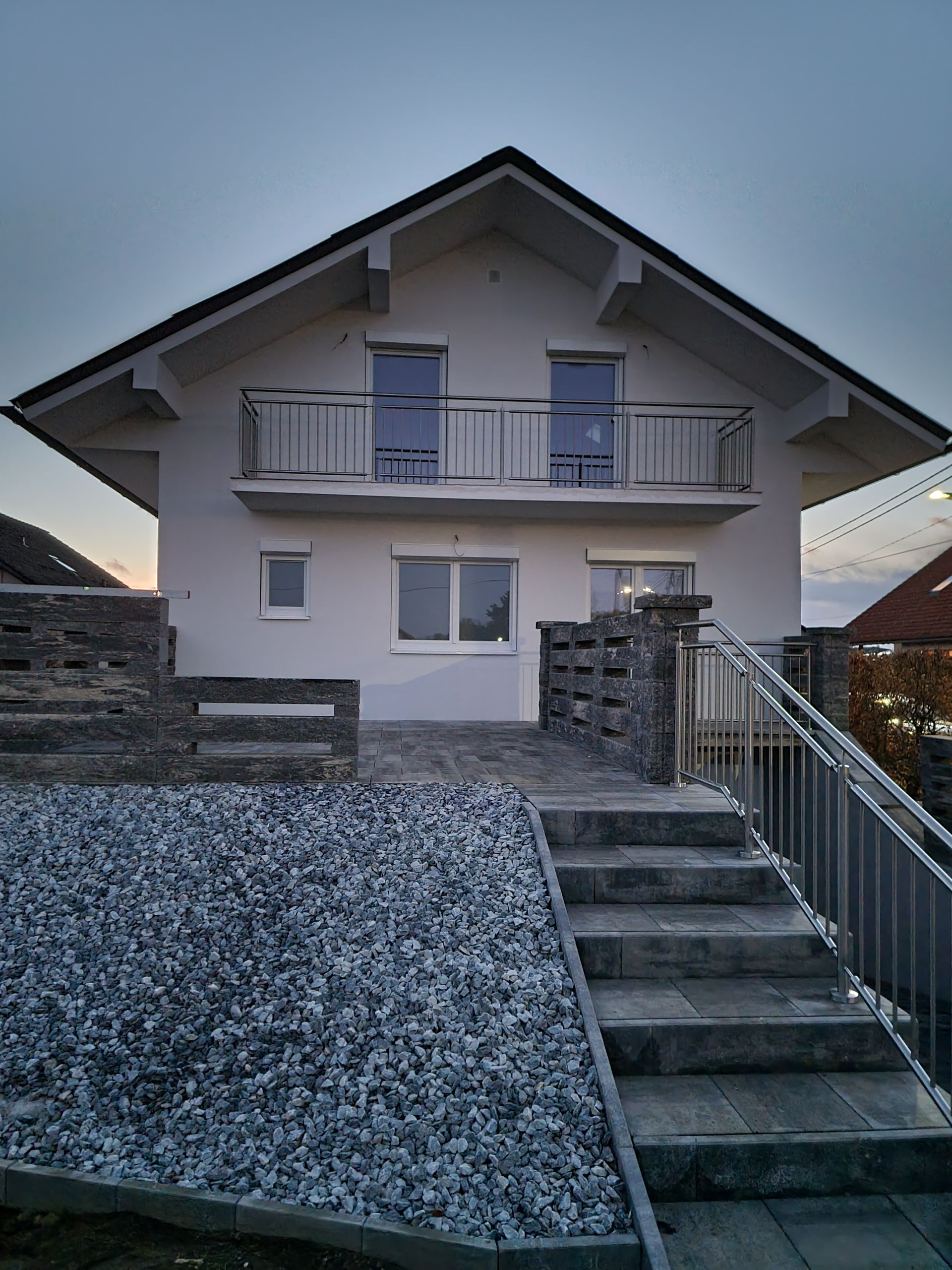 Zweistöckiges weißes Haus mit Balkon, Treppe und Kiesstützmauer; grauer Himmel.