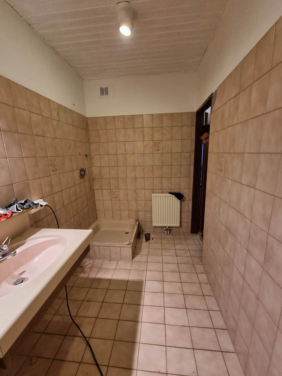 Badezimmer mit hellbraunen quadratischen Fliesen, weißem Waschbecken, Türrahmen und Heizkörper.