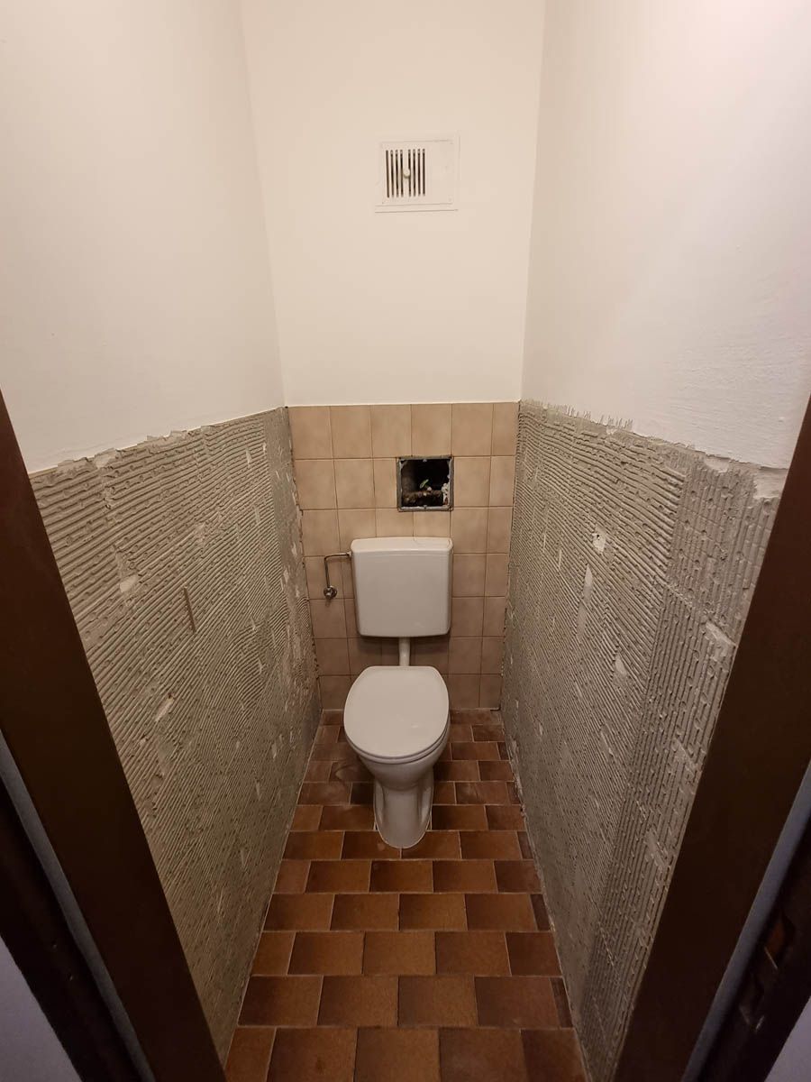 Toilette in einem kleinen, schmalen Badezimmer mit teilweise entfernten Wandfliesen und braun gefliestem Boden.