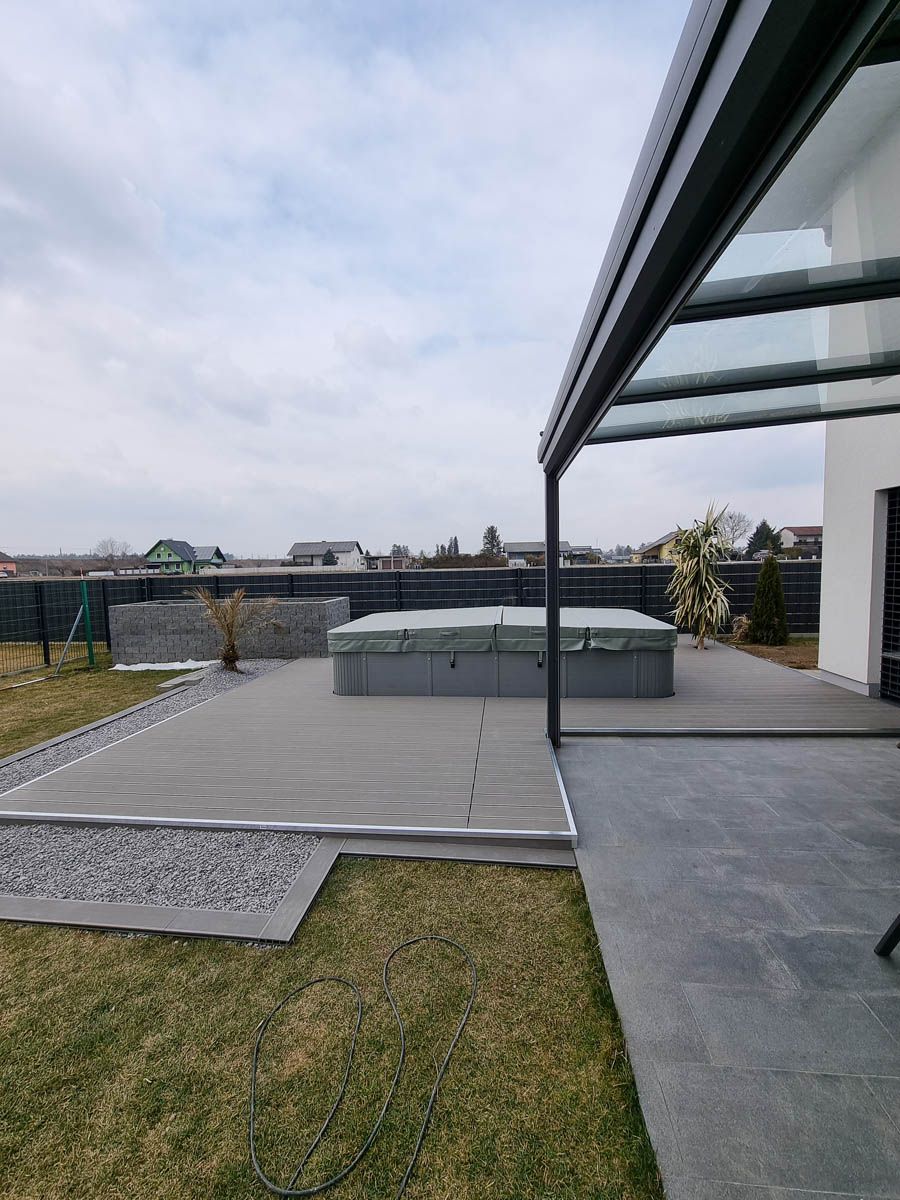 Außenterrasse mit Whirlpool unter einer Glasdachkonstruktion, grauem Deck, Rasen und bewölktem Himmel.