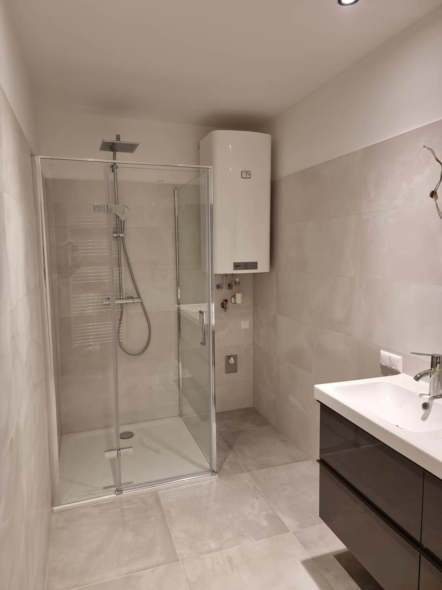 Modernes Badezimmer mit Dusche, Waschbecken und Warmwasserbereiter; hellgrau geflieste Wände und Boden.