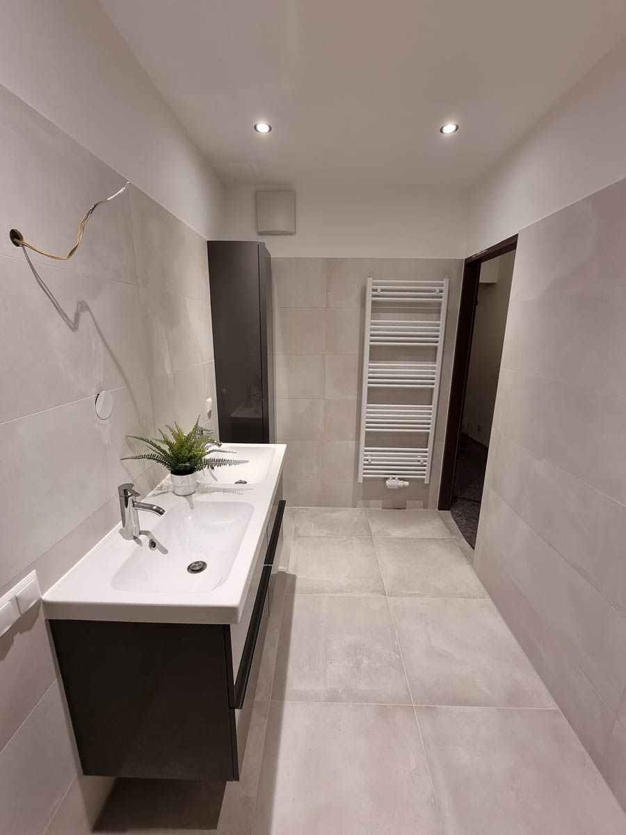 Modernes Badezimmer mit langem Waschtisch, hohem Schrank und Handtuchwärmer. Hellgraue Fliesen und Wände.