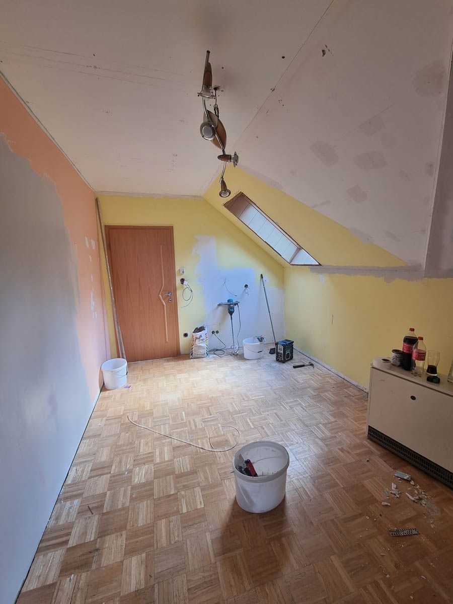 Zimmerrenovierung: Wände teilweise pfirsichfarben, gelb und weiß gestrichen. Holzboden, freiliegende Deckenleuchten. Eimer, Werkzeuge.