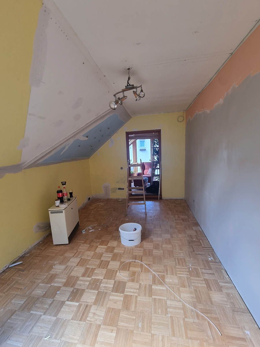 Zimmer im Umbau mit gelben Wänden, schräger Decke, Holzboden, Eimer und einem Fenster.