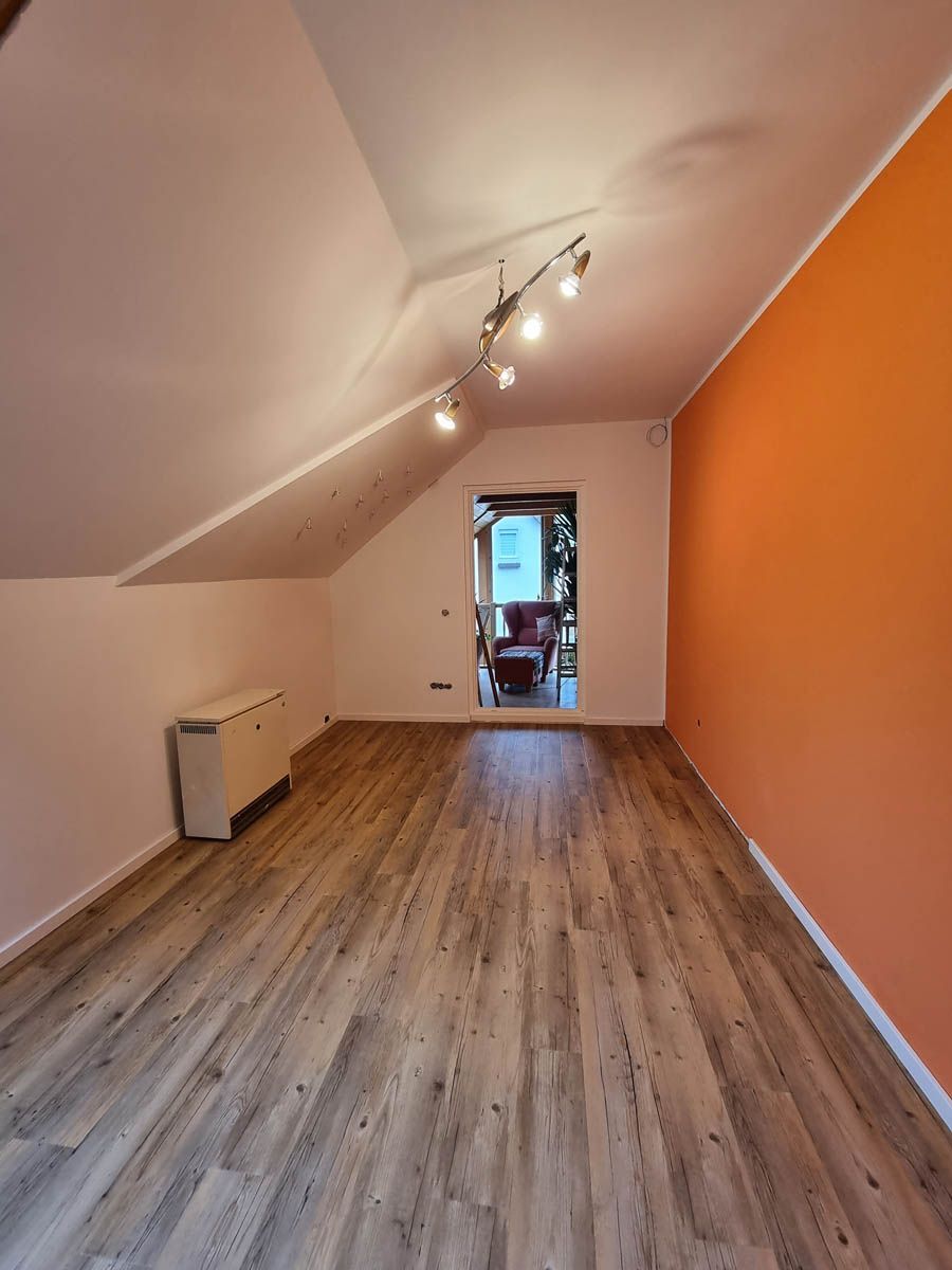 Zimmer mit Holzboden, weißen Wänden, orangefarbener Akzentwand und Deckenbeleuchtung. Durch die Tür gelangt man in ein weiteres Zimmer mit einem roten Stuhl.