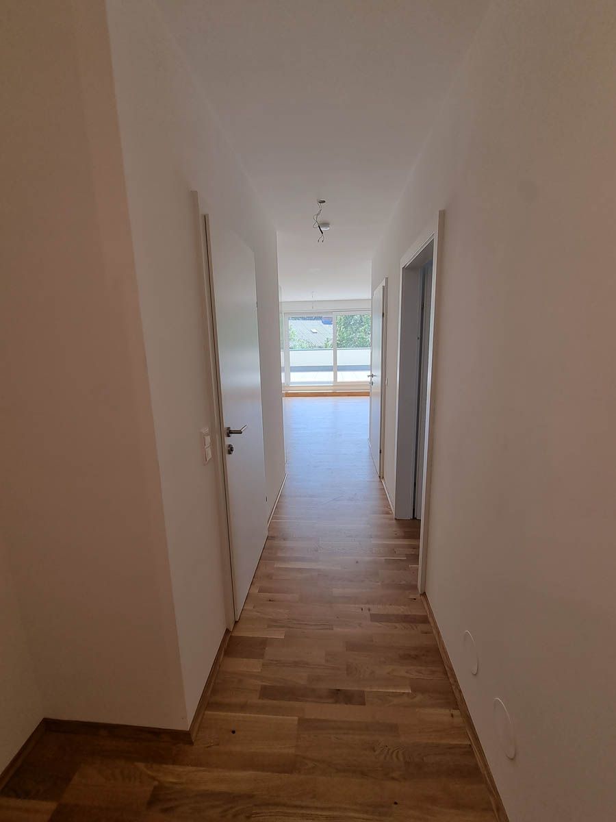 Schmaler Flur mit Holzboden, weißen Wänden und Türen, die zu einem hellen Raum mit einem großen Fenster führen.