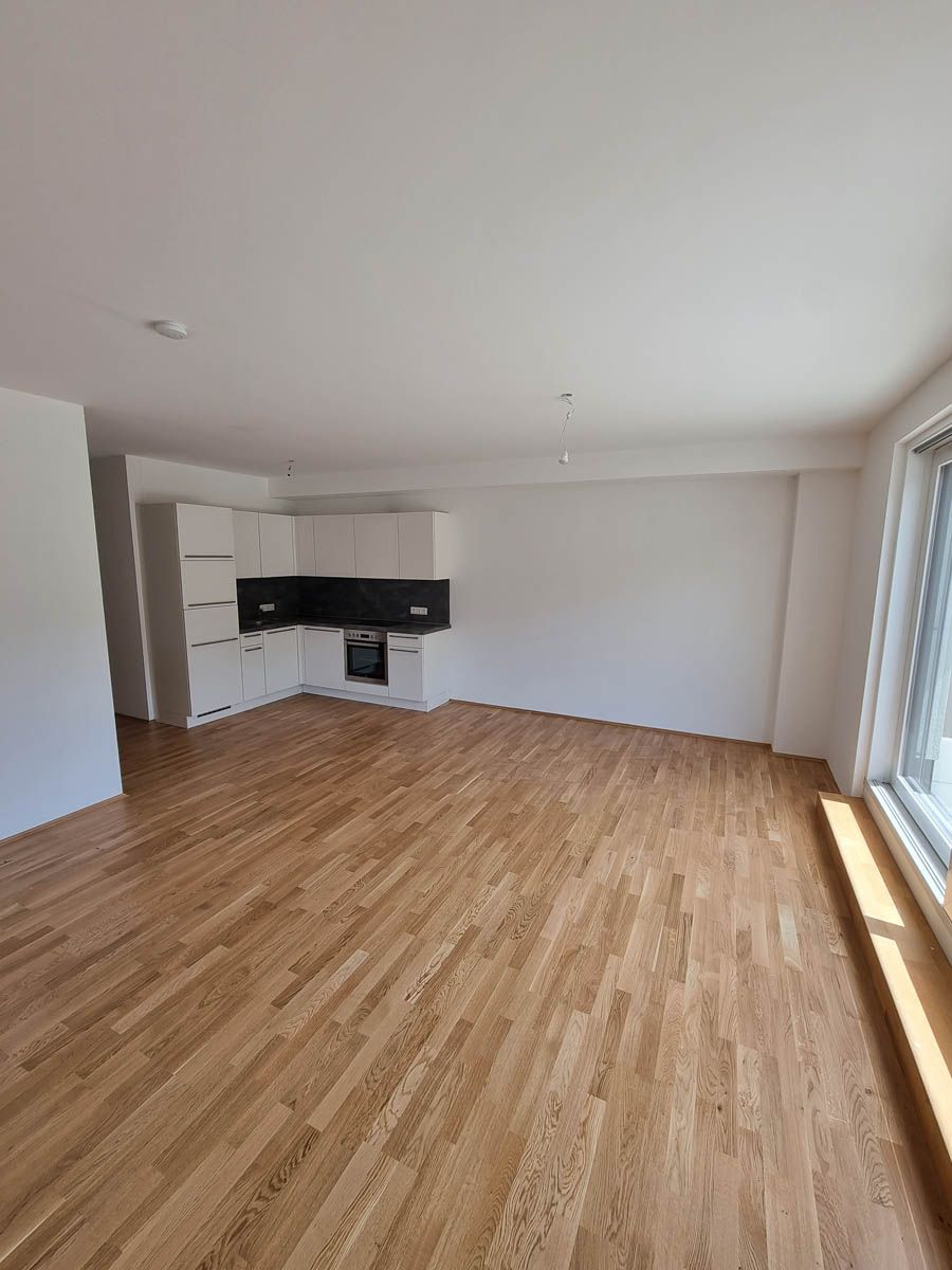 Leere Wohnung mit Hartholzböden, weißen Wänden und einem kleinen Küchenbereich. Sonnenlicht fällt durch ein Fenster.