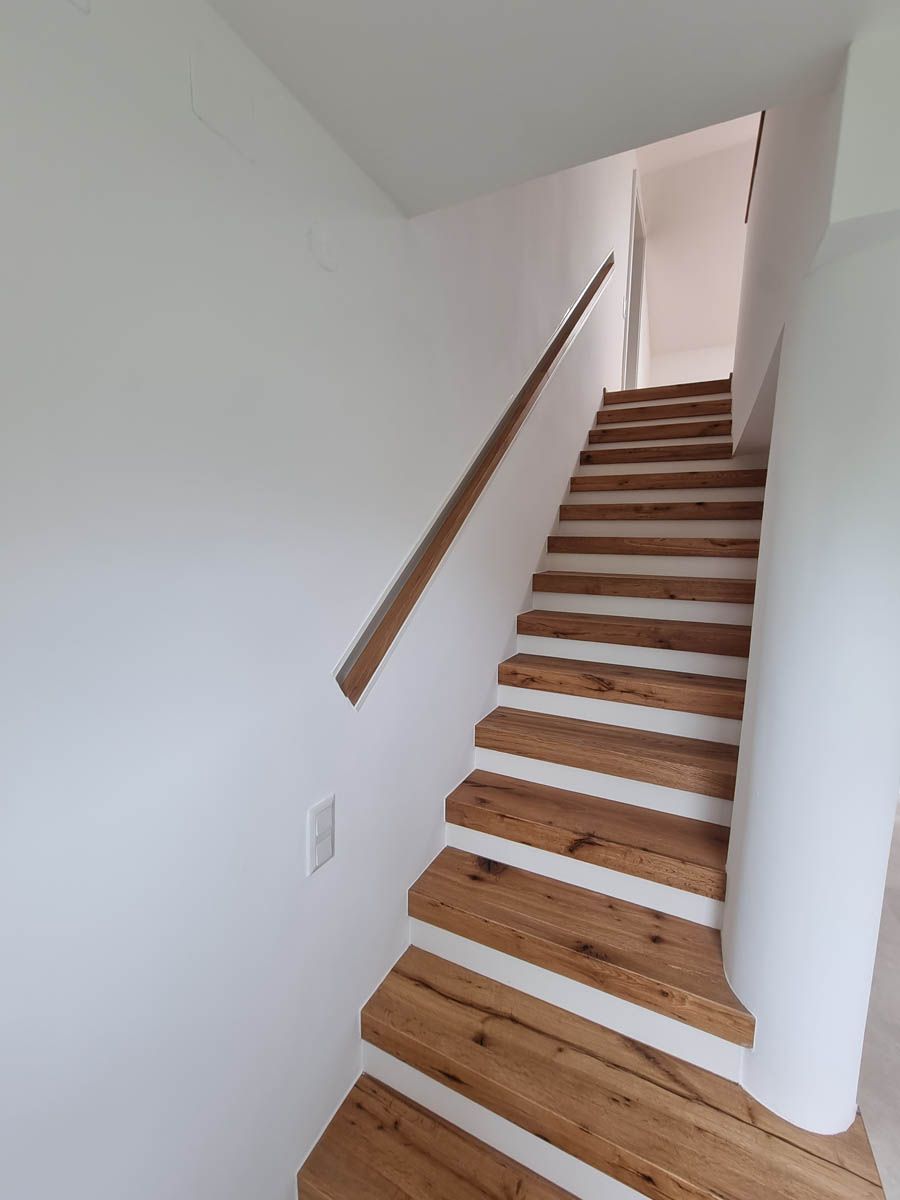 Nach oben führende Holztreppe mit geschwungenem Handlauf vor einer weißen Wand.