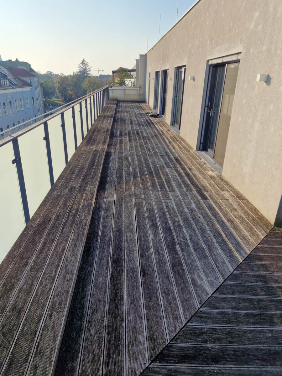 Holzterrasse auf einem Gebäudedach mit Glasgeländer. Sonnenlicht wirft Schatten.