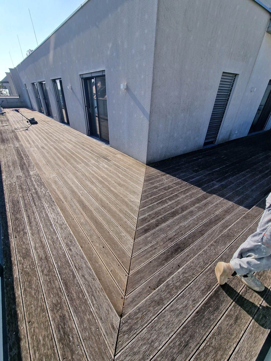 Holzterrasse an der Gebäudeecke, Person putzt sie, weiße Wand und Fenster.