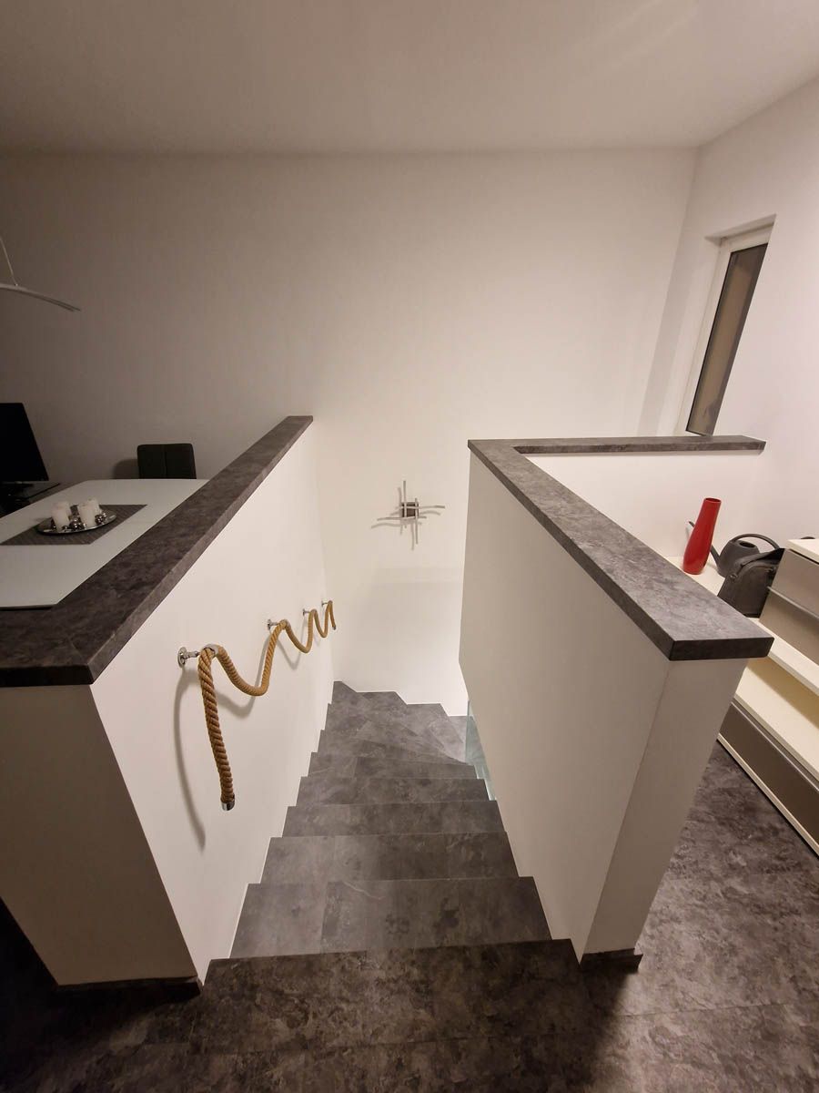 Treppe mit grauen Stufen, weißen Wänden und einem Seilhandlauf. Eine dunkle Arbeitsplatte grenzt an die Wände.