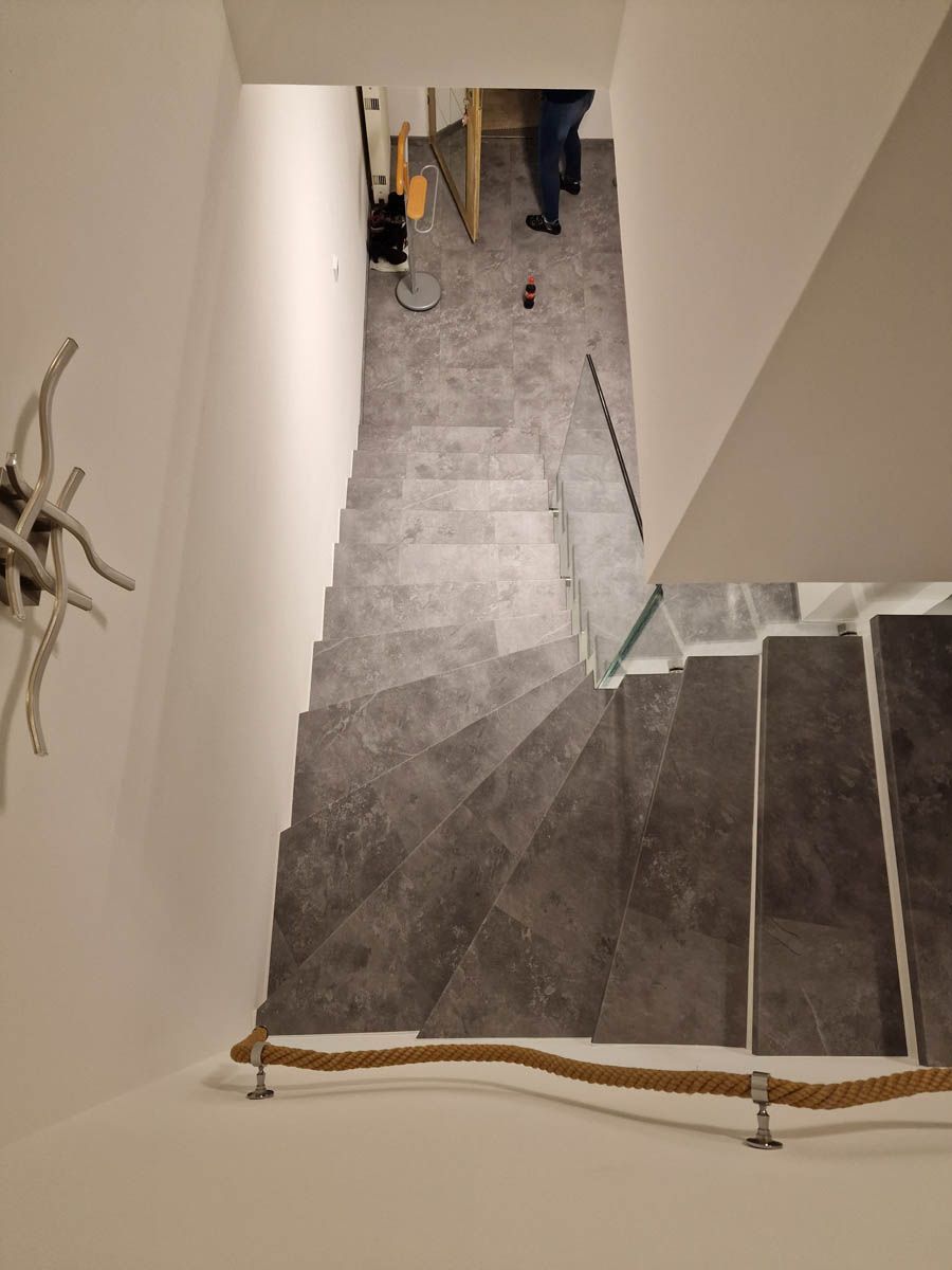 Treppe mit grau gefliesten Stufen, Seilhandlauf und einer Person oben. Weiße Wände.