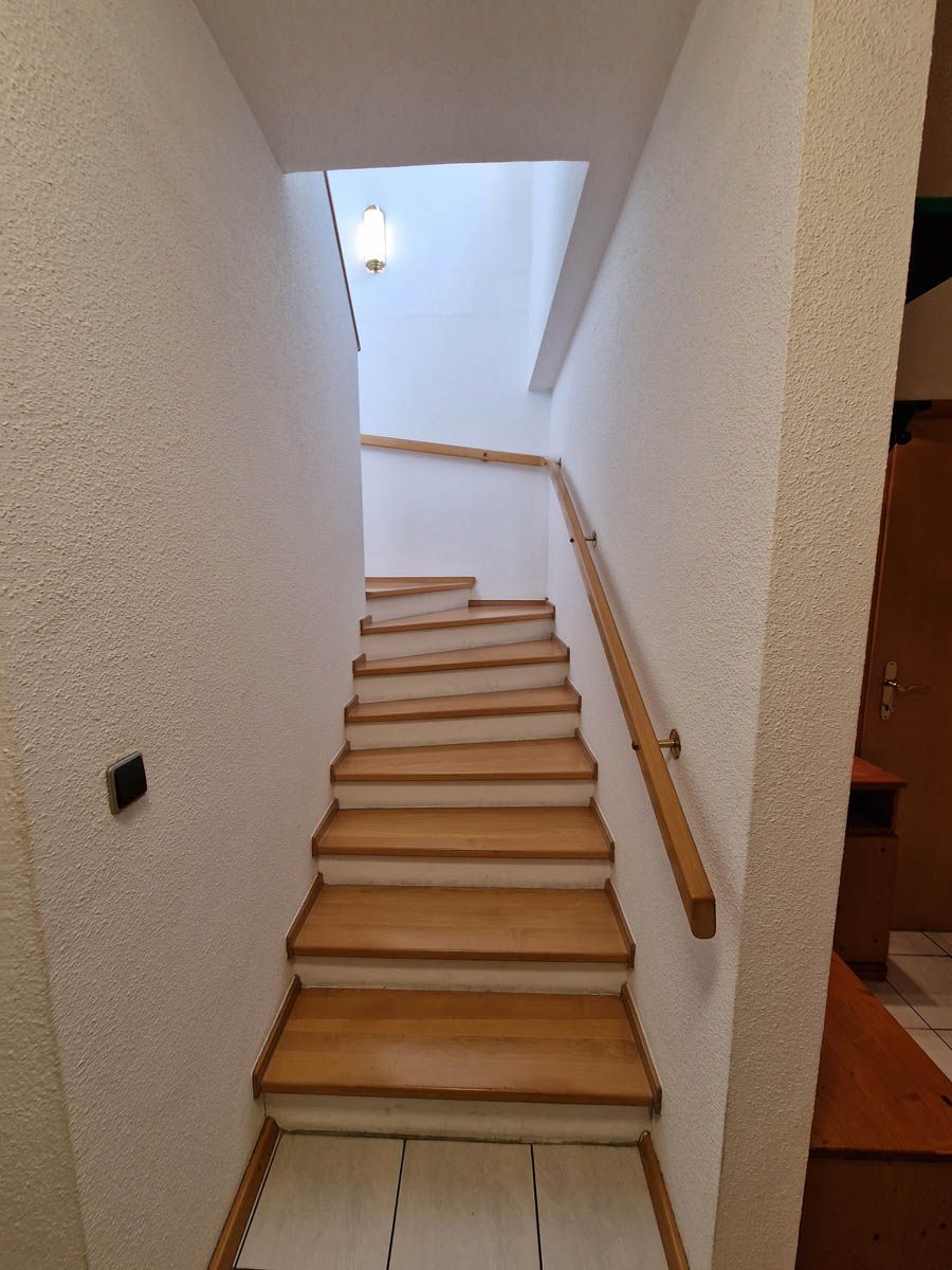 Treppe mit Holzstufen und Handlauf nach oben. Weiße strukturierte Wände.