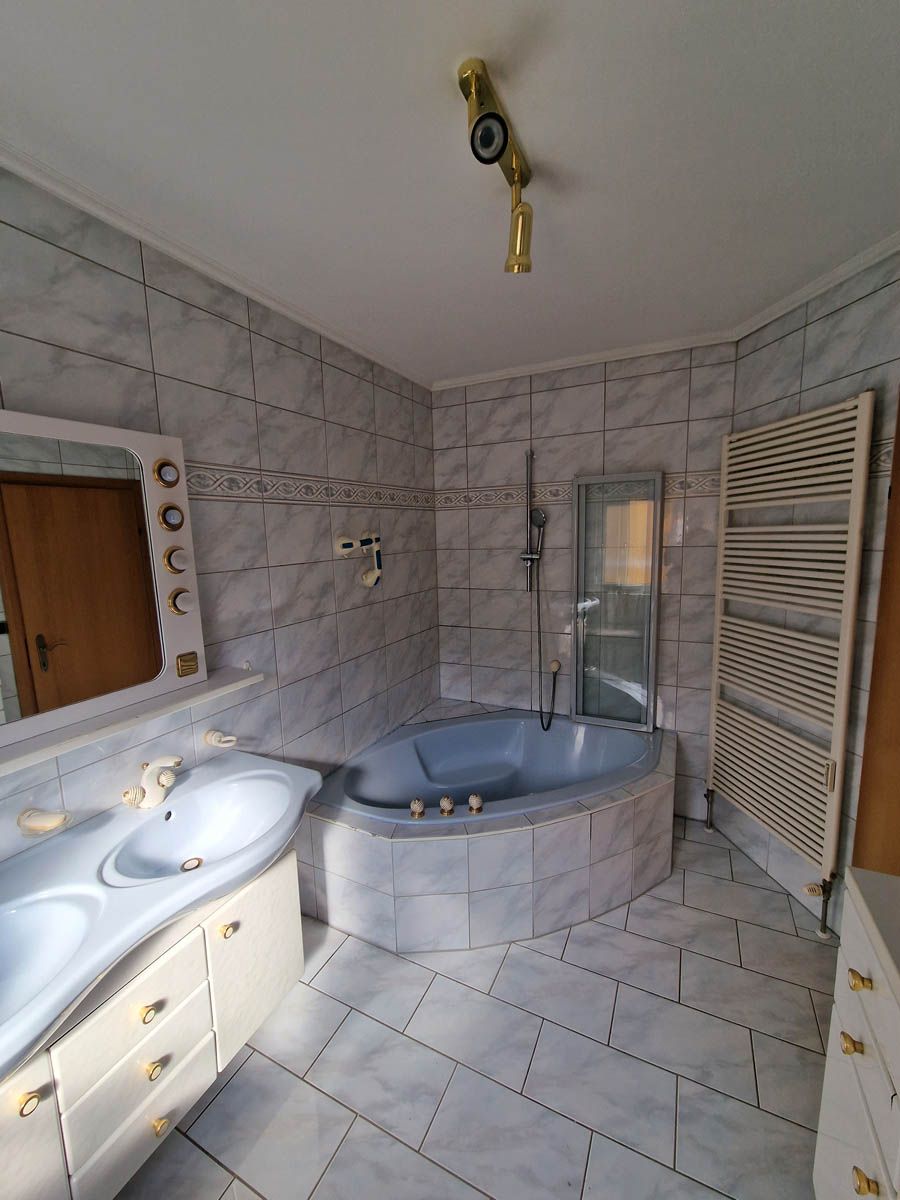 Badezimmer mit grau gefliesten Wänden und Boden, Eckbadewanne, Doppelwaschbecken und Handtuchwärmer.