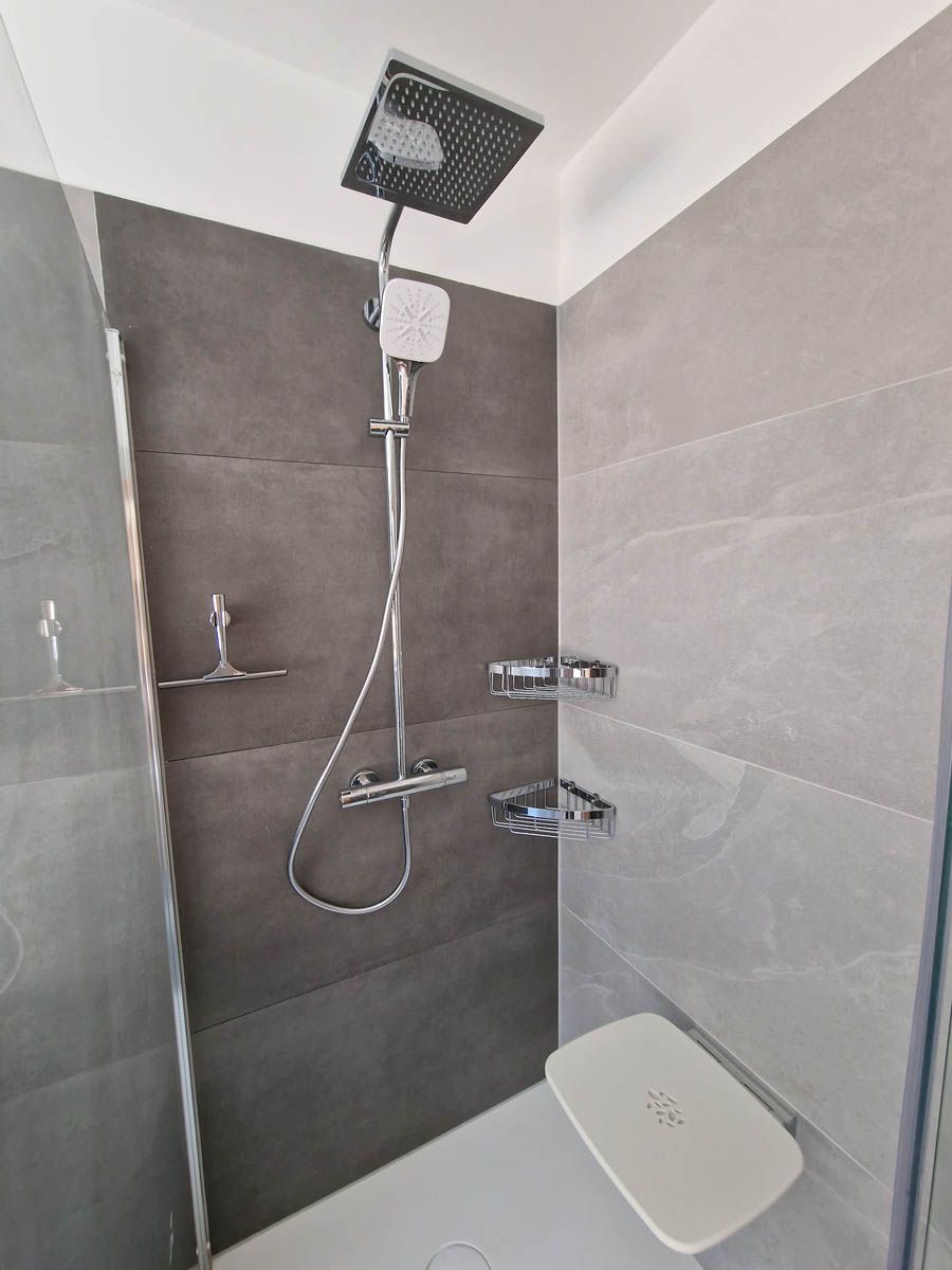 Duschkabine mit grauen Fliesen, Chromarmaturen, Duschkopf und Klappsitz.